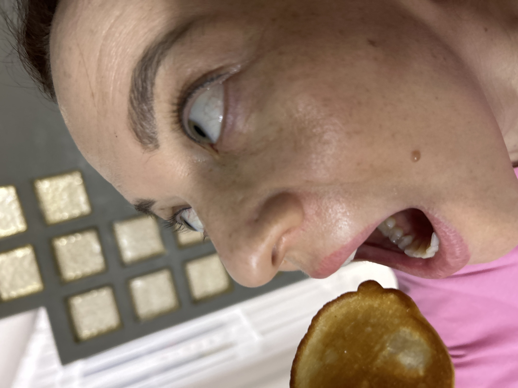 A close-up, low-angle shot shows a person with pale skin, an open mouth, and eyes looking upward. The person is about to eat a golden-brown object that resembles a pancake or pastry. The person's face is turned to the side, revealing the nose, cheek, eyebrow, and teeth. A mole is visible on the cheek. The background contains a pattern of small squares that appear to be tiles or containers. A white surface is visible beneath the object being eaten. The person is wearing a pink garment. The location is not provided in the image and therefore cannot be determined.