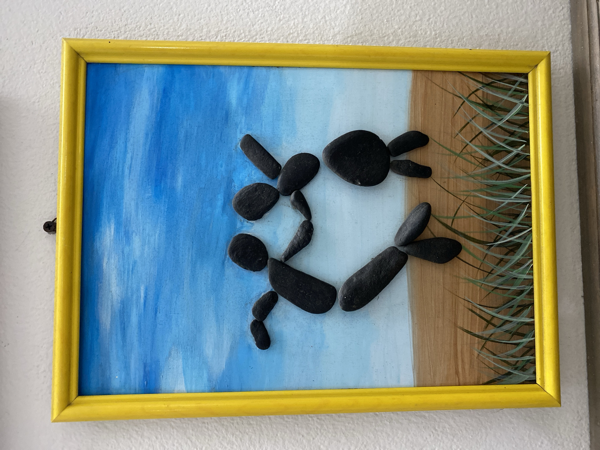 This is a framed piece of art depicting two stick figures made of black stones dancing. The background is a painted sky and a field of grass. The art is hung on a white textured wall. A yellow frame surrounds the artwork.