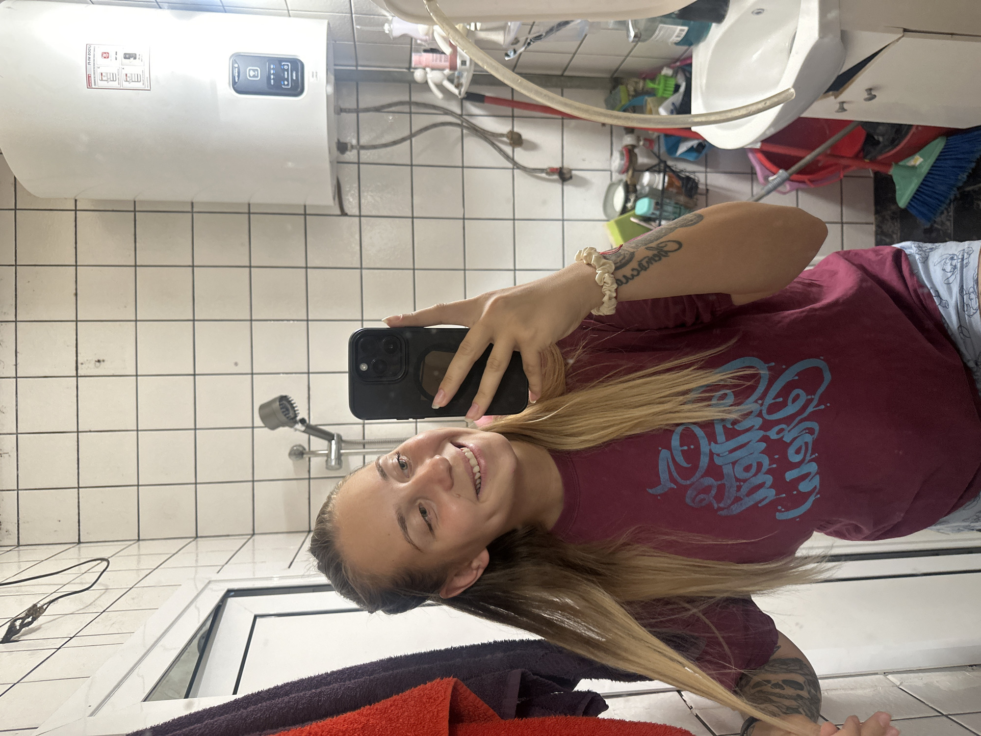 In a bathroom, a young woman with blonde hair, wearing a maroon t-shirt and light-colored shorts, takes a mirror selfie. She holds a black smartphone in her right hand, which is adorned with a scrunchie bracelet. Her left arm, featuring a tattoo, is extended as she touches her hair. The bathroom walls are covered in white square tiles. To the left, a red and a purple towel hang on a rack. A water heater is mounted on the right wall. In the background, a sink is visible, along with cleaning supplies and a broom. The scene appears to be set in a residential apartment bathroom in the city of Plovdiv, Bulgaria.