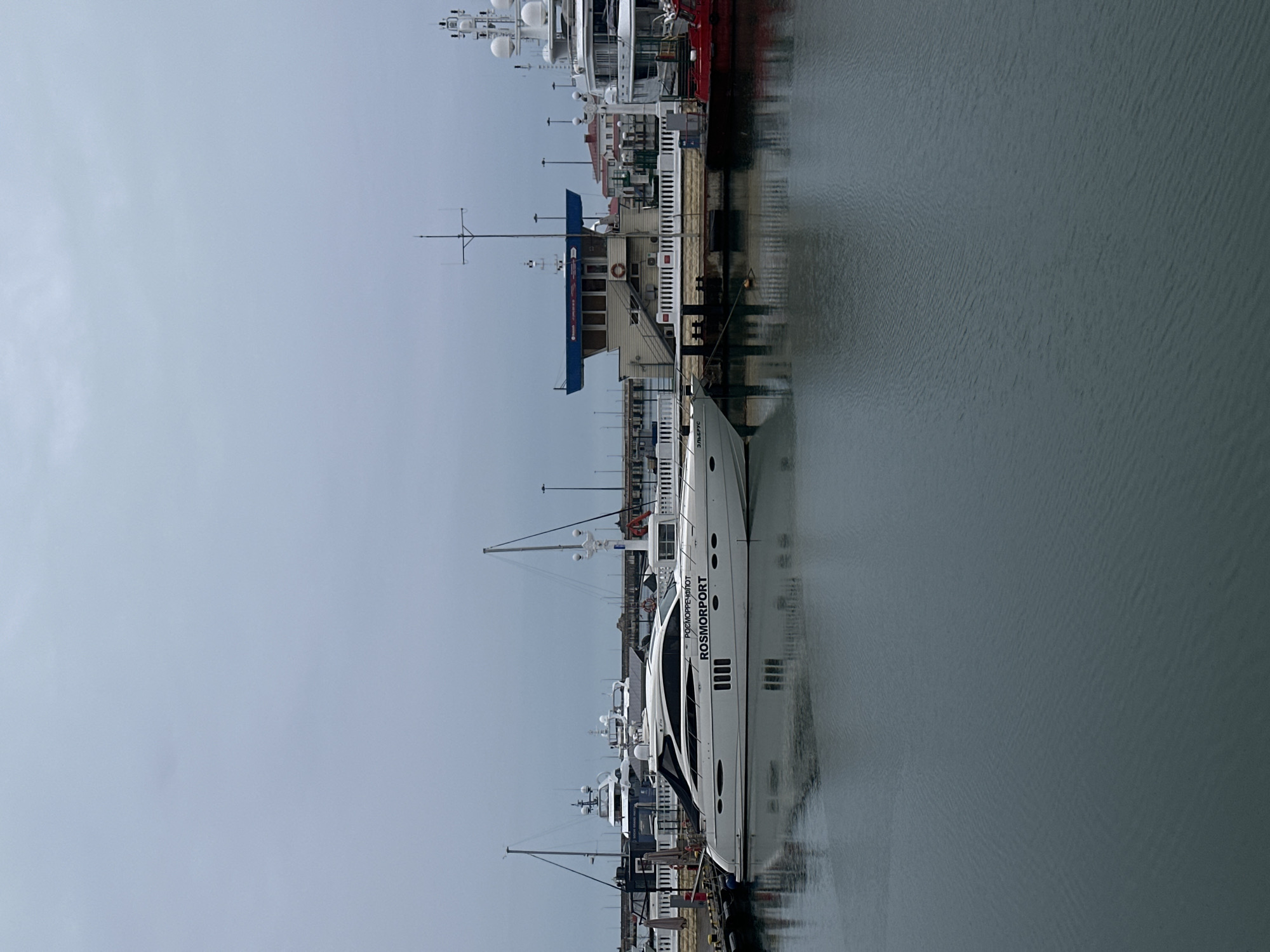 A sideways shot depicts a harbor scene, possibly in Sochi, Russia, given the Russian language on one of the boats. Several yachts and boats are docked along the harbor wall. The water is still, with a slight ripple effect. The sky is overcast and gray. A building with a blue roof is present, suggesting a maritime-related structure or office. Street lamps are visible along the harbor side, further indicating this is an urban setting. The overall scene suggests a calm, perhaps inactive, day at the harbor.