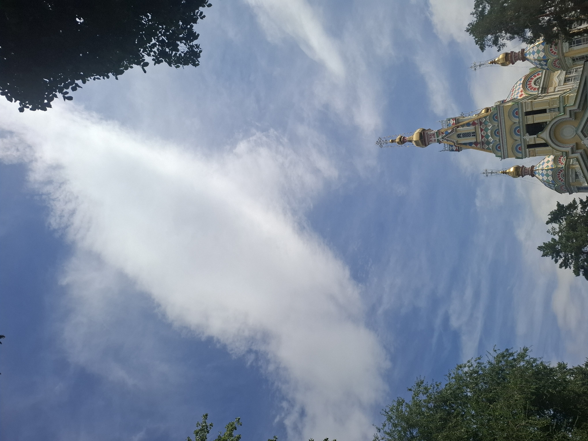 The image shows a low-angle view of Zenkov Cathedral, also known as Ascension Cathedral, in Almaty, Kazakhstan, against a backdrop of a blue sky with scattered white clouds. The cathedral's ornate golden domes and colorful tiled roof are visible, along with some of the surrounding trees. The lighting suggests it is daytime. There are no people or animals in the frame. The image appears to be taken from a public outdoor space.