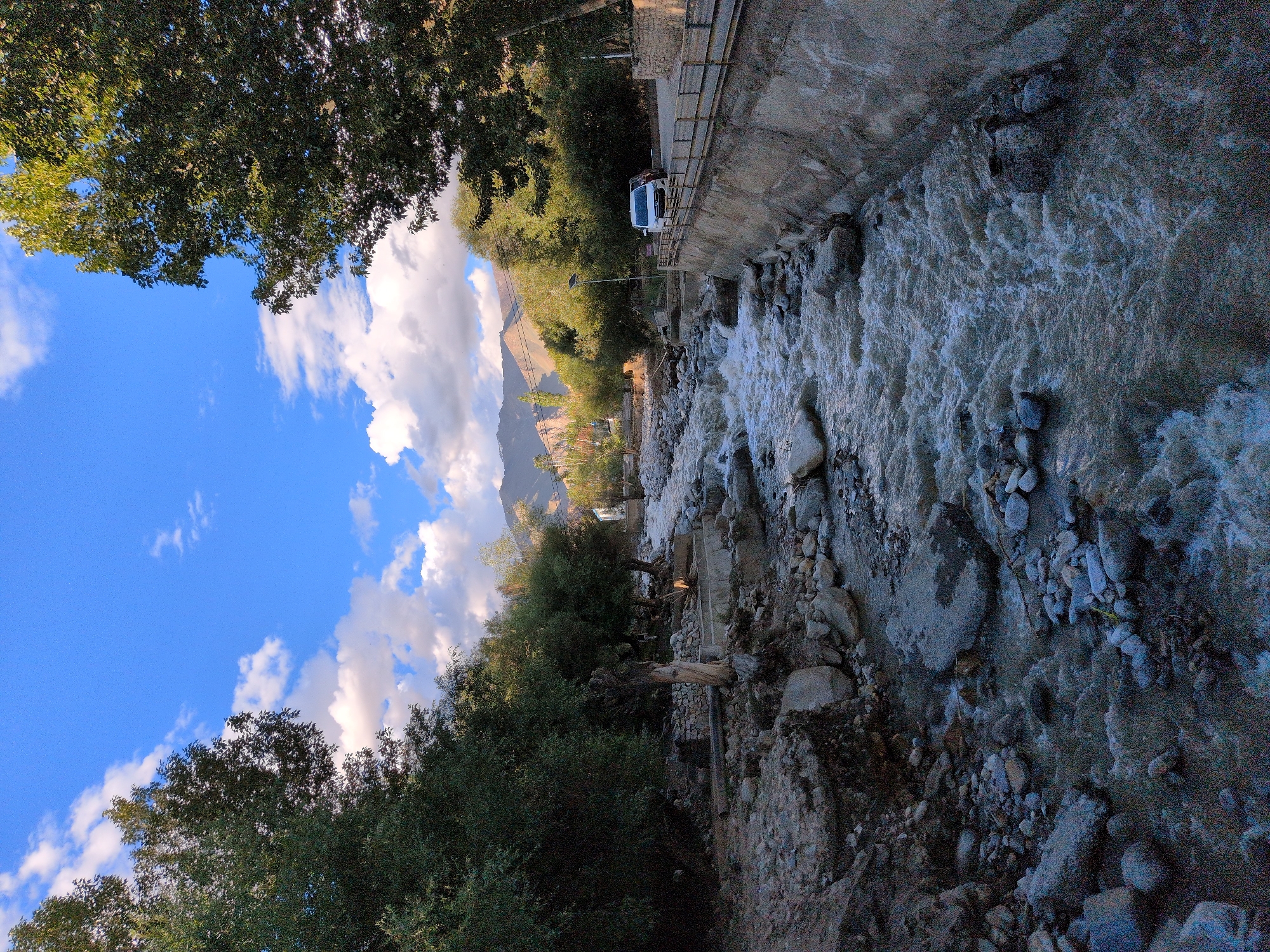 The image shows a fast-flowing river in Leh, India, with rocky banks and trees lining the sides. A white van and a red car are parked on the side of the road next to a concrete retaining wall. Mountains are visible in the background under a blue sky with white clouds.