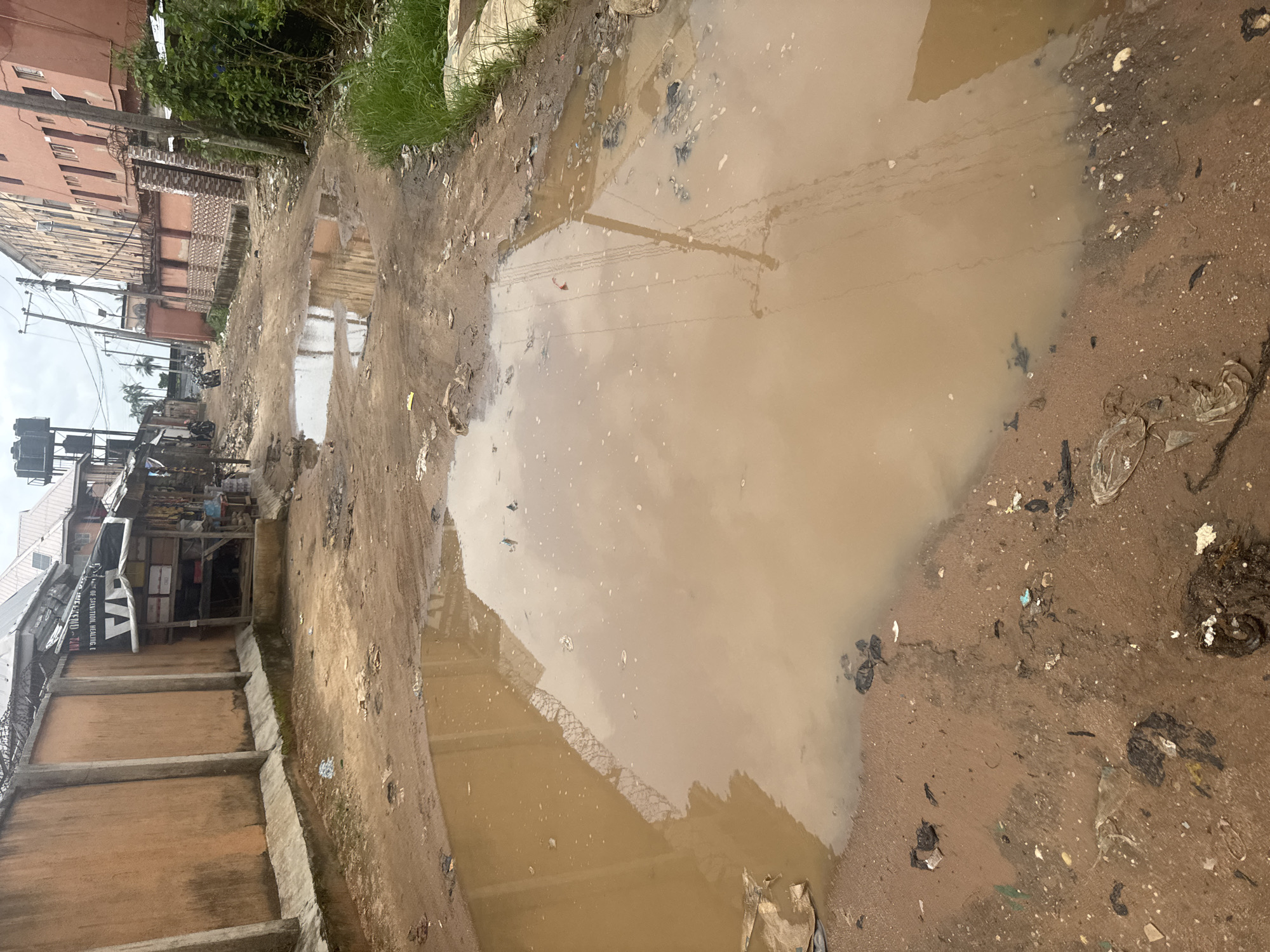 This image depicts a muddy, flooded area in Ihiagwa, Nigeria, likely a street or pathway. The ground is covered in brown mud and rainwater, forming a large puddle that reflects the overcast sky and some overhead wires. Debris, including plastic bags and other refuse, is scattered around the edges of the puddle and on the surrounding dirt.
To the left of the puddle, there is a row of buildings. The closest building appears to be a multi-story structure with a light pink facade. Further down, there are smaller structures, possibly shops or residences, with awnings and open fronts displaying various items. A black and white banner with text is visible on one of these structures. A car with "TAX" written on its side is parked near the shops. A tall metal pole with wires stretches across the scene, with its reflection visible in the water.
Vegetation, including grass and some leafy plants, grows along the edge of the buildings and the pathway. The overall scene suggests an urban or semi-urban environment that has experienced recent rainfall, leading to the accumulation of water and mud. The lighting indicates it is daytime, with diffused light suggesting cloudy weather. There are no people or animals clearly visible in the frame.
The visible text on the banner reads: "OF SANITATION HEALTH & ... TAXI".