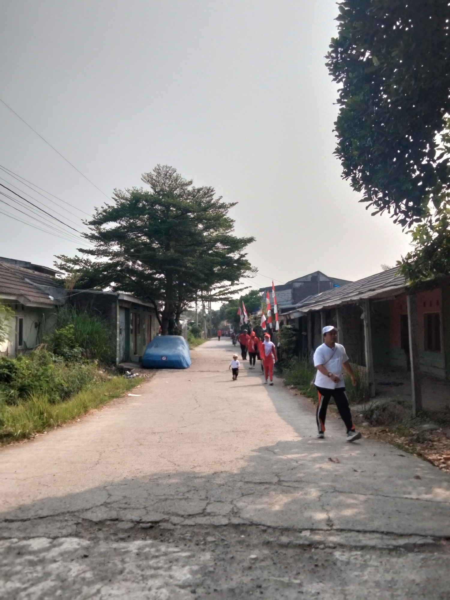 In Cariu, Indonesia, an outdoor scene captures a group of people walking along a cracked asphalt road lined with modest houses and lush vegetation. The sky is overcast with a pale haze. On the left, a large tree with dense foliage stands near a house with a gray tiled roof and white walls. A blue tarp covers a vehicle parked beside a row of buildings. In the distance, a group of individuals, some wearing red and white attire, walks together, accompanied by Indonesian flags. A young child is also part of the group. Closer to the foreground on the right, a man in a white t-shirt and dark pants walks toward the viewer. The road exhibits visible wear and tear, with numerous cracks and patches.