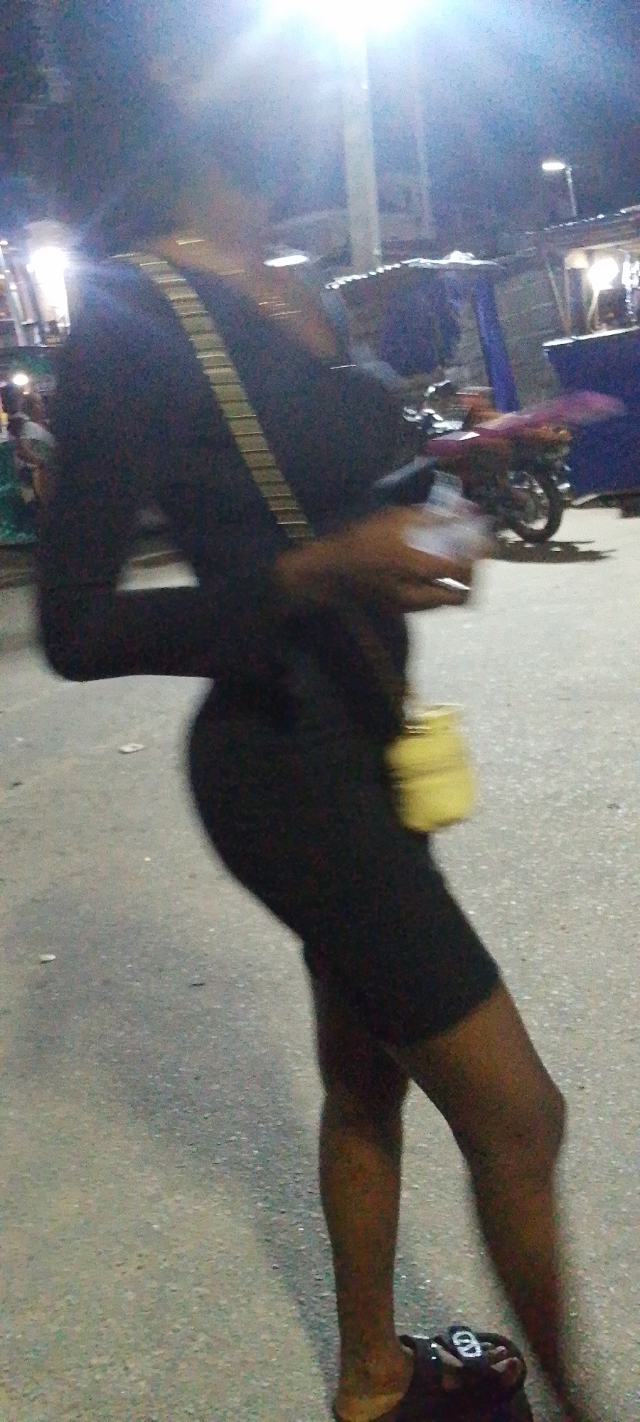 A person stands on a street in Lokoja, Nigeria. The individual wears a dark, long-sleeved dress or jumpsuit and a crossbody bag. A motorcycle is parked nearby. The location appears to be an urban area at night, possibly with street vendors present.