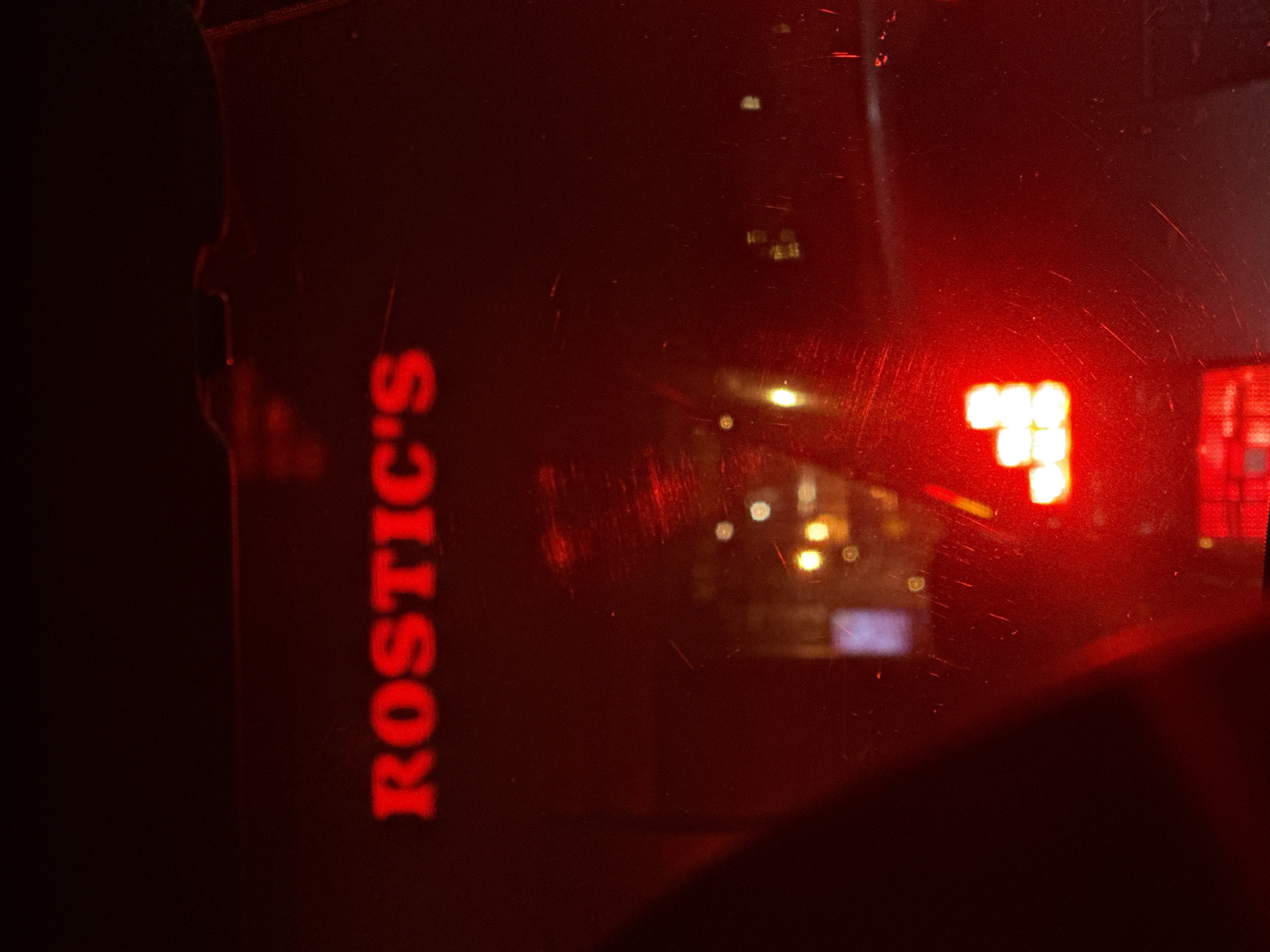 The image, taken from within a vehicle at night, depicts reflections of external objects on the car window. On the left, the vertical, red neon sign "ROSTIC'S" is reflected. In the center, blurred street lights are visible, while on the right, a red light display, possibly a car taillight. The image is dark, and the reflections create a distorted view of the outside environment. While "ROSTIC'S" is a fast-food chain found in Krasnoyarsk, Russia, the image lacks sufficient details to confirm a specific location within the city. A portion of a person's silhouette appears on the left edge of the image, near the Rostic's sign.