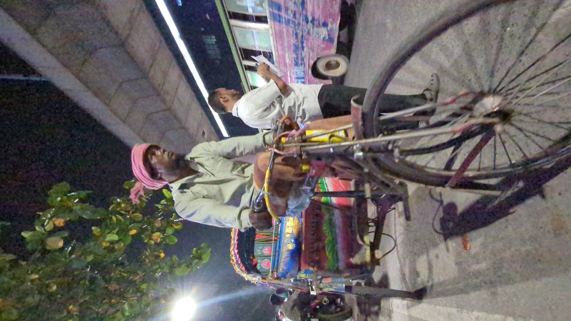 The image depicts a scene in Dhaka, Bangladesh at night. A rickshaw puller wearing a pink turban and light green shirt sits on his colorfully decorated rickshaw. Another man stands near a bus, appearing to read something. The scene is illuminated by streetlights and lights from the bus. A tree with green leaves and small yellow fruits is visible on the left side of the frame. An elevated concrete structure is visible in the background. The scene suggests a street-level interaction in a busy urban environment.