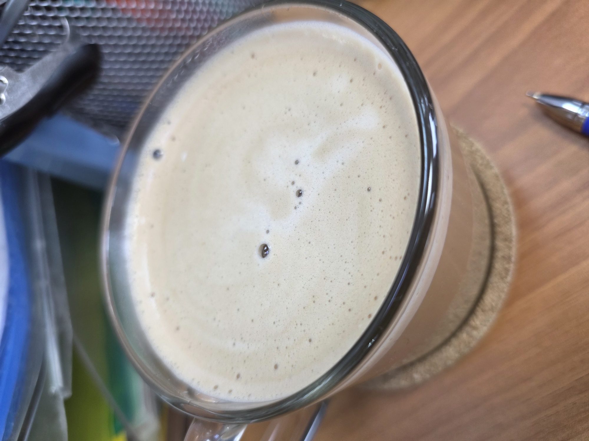 A close-up, high-angle shot of a clear glass mug filled with a light-brown, frothy liquid (likely coffee or latte) resting on a round, beige coaster. The coaster and mug are situated on a light-brown wooden surface. In the background, to the left, is a blurred collection of items including a black object (possibly a clip) and various blue, green and yellow items within a container. To the right, a portion of a blue and silver pen is visible on the wooden surface. The image provides no evidence to confirm any location, in Chaikovsky, Russia or anywhere else.