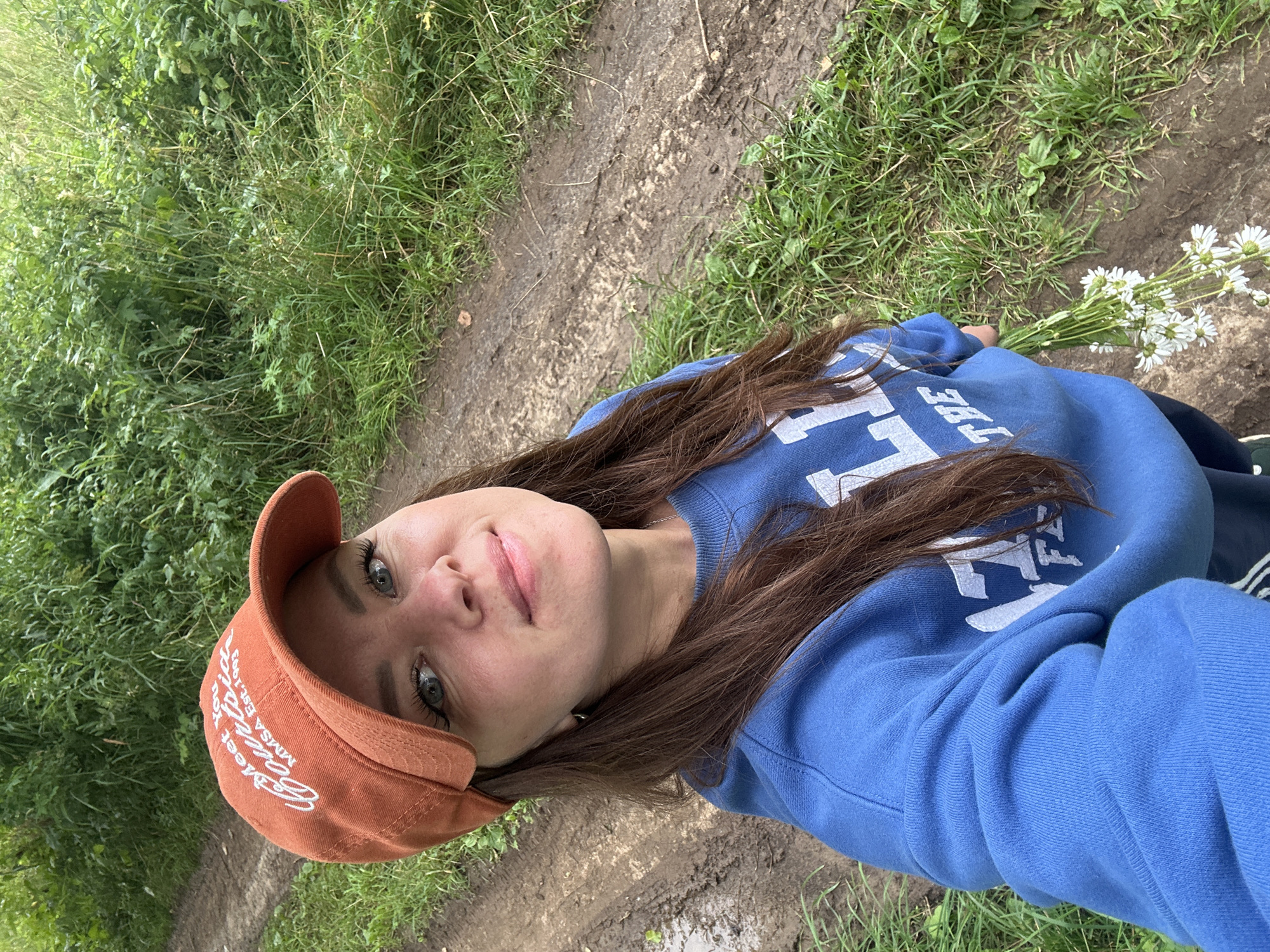 A woman with long brown hair, wearing an orange baseball cap and a blue sweatshirt, takes a selfie on a dirt path surrounded by green foliage. The cap has white text that reads "Meet You Courtside MMSA Est. 1993". She is holding a small bouquet of white flowers in her right hand. The path appears to be in a rural or natural setting, possibly a park or wooded area, and is wet with mud. The background is filled with lush green grass and plants. The image was taken outdoors, likely during the daytime, with natural lighting. The location in the city of Noviy, Russia is not discernible from the image itself.