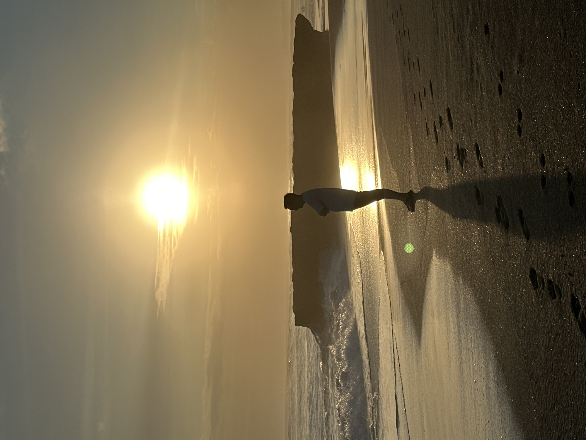 An individual stands on a sandy beach, silhouetted against a bright, setting sun. The sun, positioned high in the sky, casts a warm, golden glow over the scene, with the light reflecting off the wet sand and the gentle waves. In the distance, a rocky outcrop juts out into the ocean, its dark form contrasting with the illuminated water. Footprints are visible in the sand, indicating the person's path. The scene appears to be the beach in North Kuta, Indonesia, during sunset.