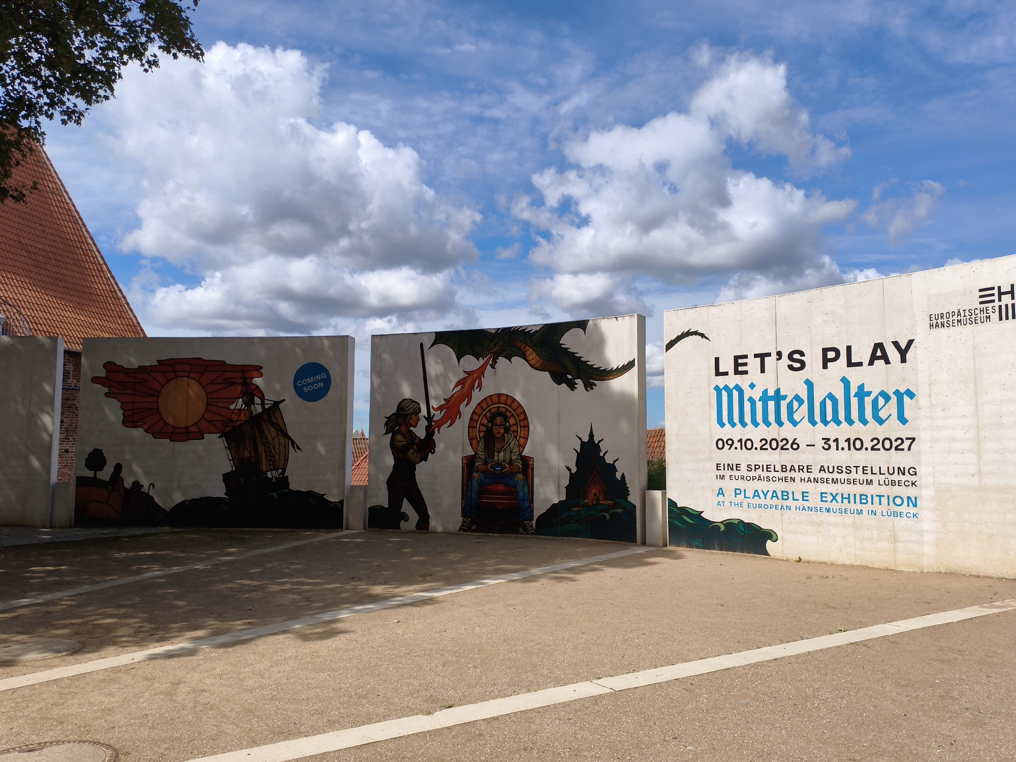 The image shows a mural on three concrete walls outside the European Hansemuseum in Lübeck, Germany. The mural depicts scenes from the Middle Ages, including a sailing ship with a large sun, a warrior fighting a dragon, and a person sitting on a throne playing a video game. The rightmost wall has text that reads "LET'S PLAY Mittelalter 09.10.2026 - 31.10.2027 EINE SPIELBARE AUSSTELLUNG IM EUROPÄISCHEN HANSEMUSEUM LÜBECK A PLAYABLE EXHIBITION AT THE EUROPEAN HANSEMUSEUM IN LÜBECK". The sky is blue with white clouds, and a tree branch is visible in the top left corner. The ground in front of the walls is paved with gravel and lined with white concrete strips.