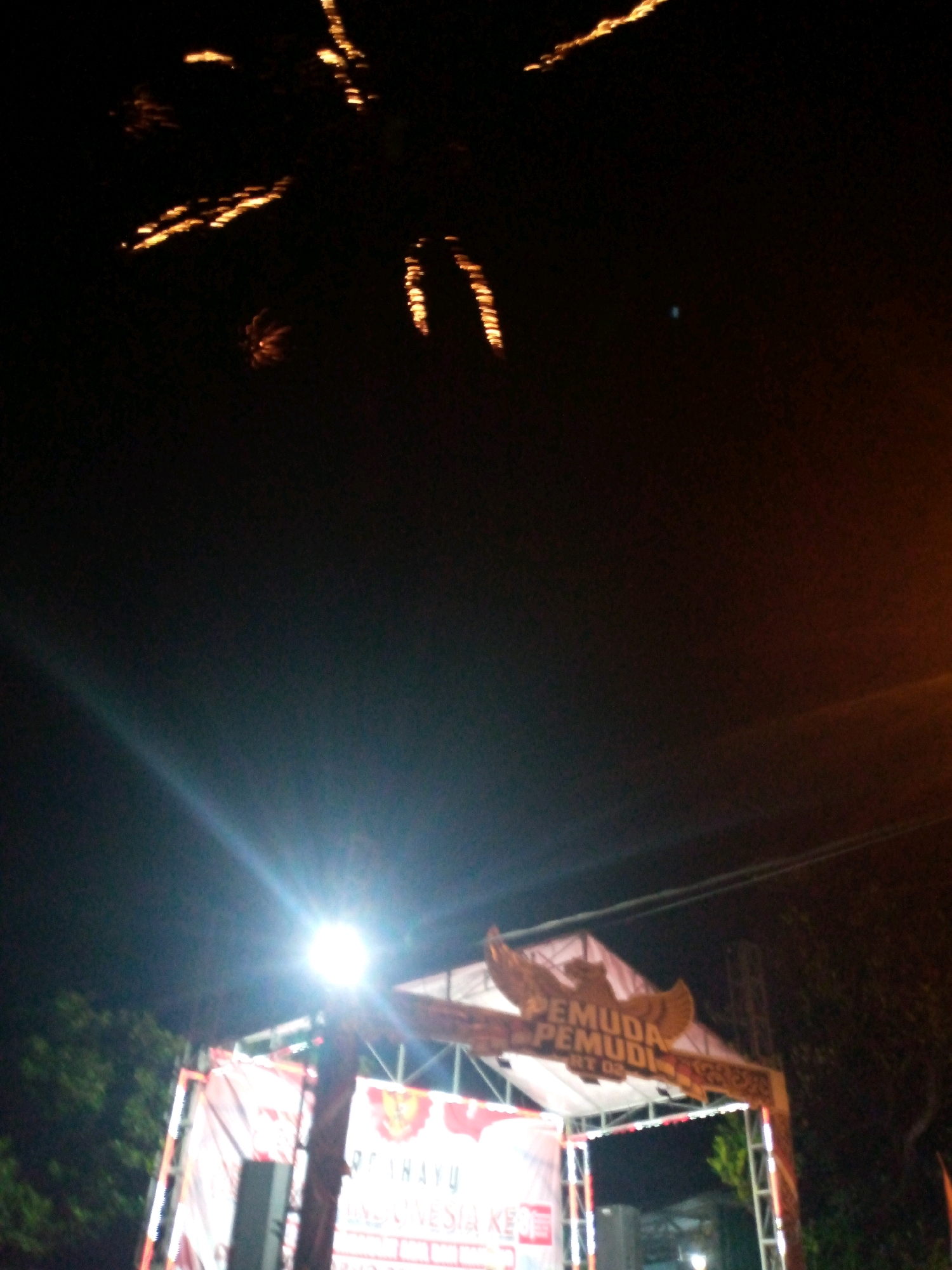 This image captures a nighttime scene with fireworks illuminating the dark sky. In the foreground, a stage or event structure is visible, decorated with banners and lights. One banner prominently displays text that appears to be in Indonesian, with a stylized eagle emblem above it. The overall atmosphere suggests a festive event, possibly a celebration or festival, taking place in Sumberwono, Indonesia. The low-light conditions and the movement of the fireworks contribute to a somewhat blurred and dynamic visual effect.