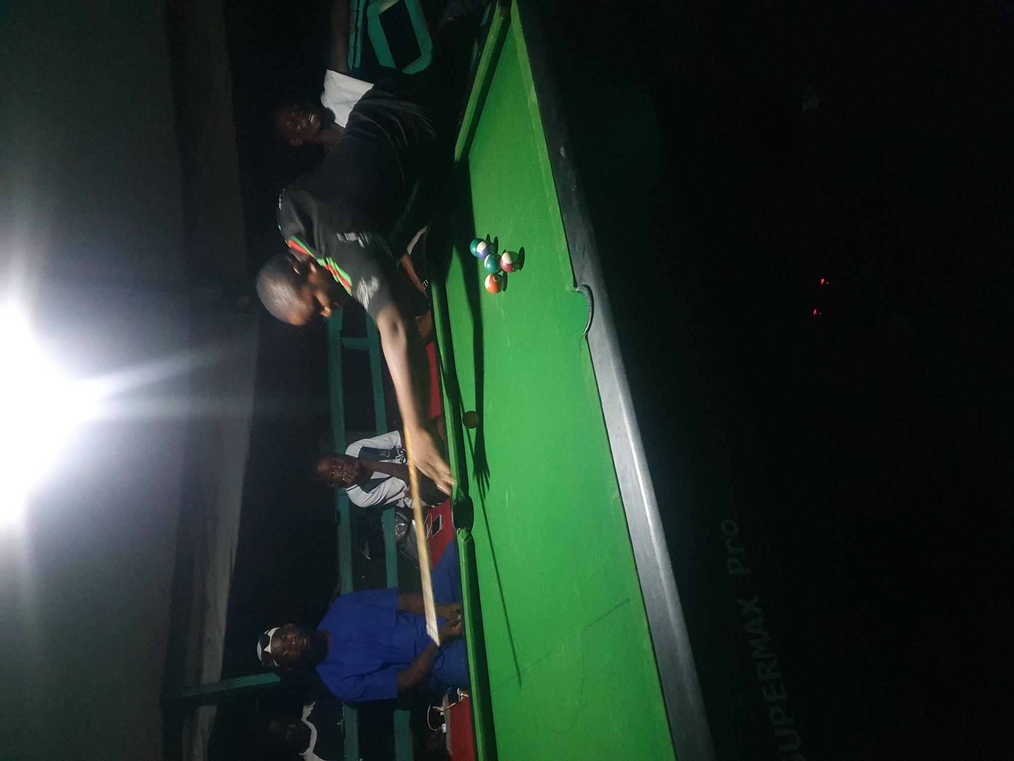 The image depicts an indoor pool hall, possibly in Lokoja, Nigeria, at night. A group of men are gathered around a green pool table. One man, wearing a black shirt with green and red accents, is actively playing pool, leaning over to strike a ball with a cue stick. Other men are seated or standing nearby, observing the game. The lighting is dim, with a bright light source visible at the top of the frame casting a glare. The pool table has several balls on it, including a cue ball, a red ball, a green striped ball, and a white ball. The word "SUPERMAX Pro" is visible on the side of the pool table. The overall atmosphere suggests a casual gathering for recreation.