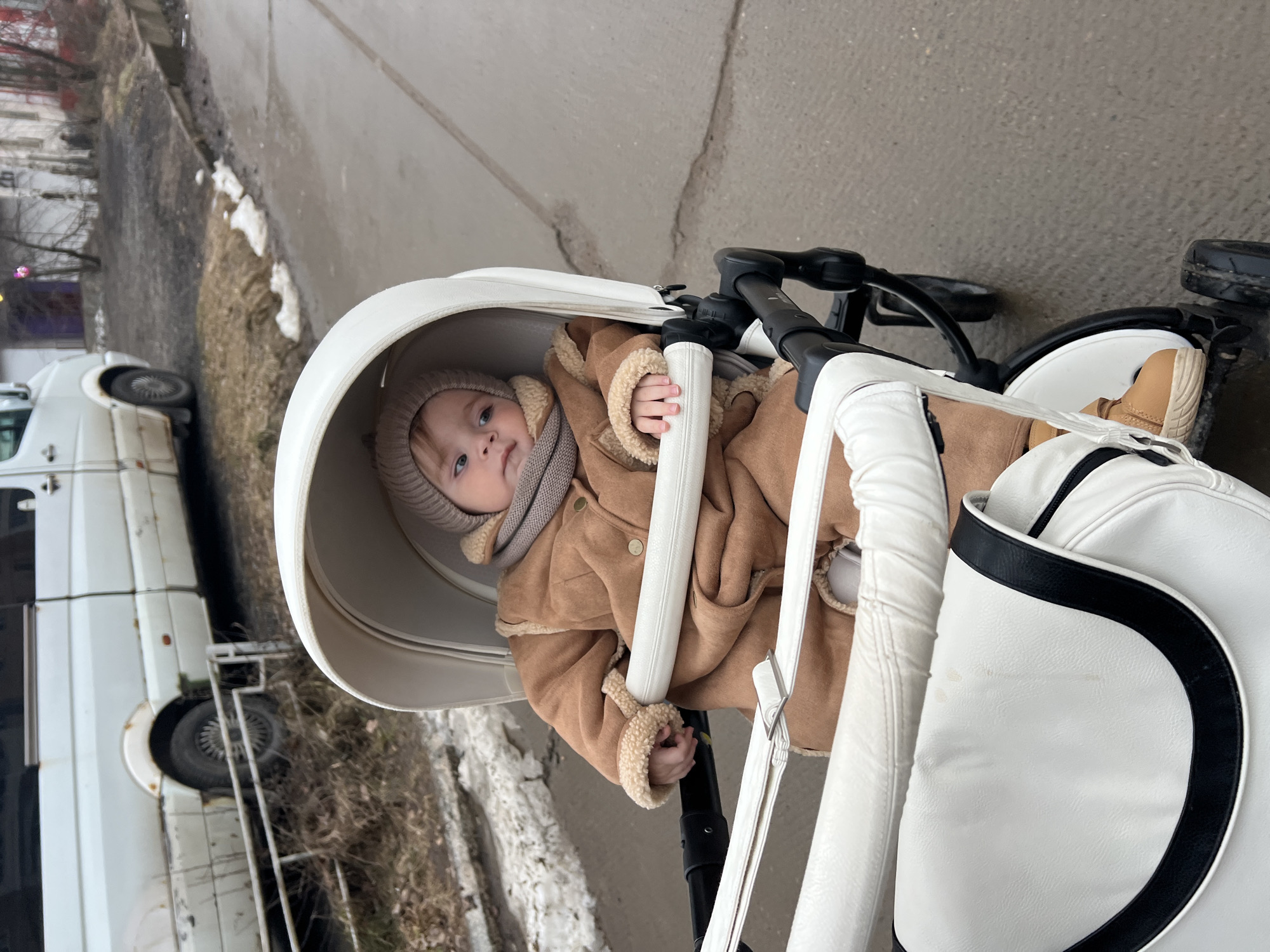 A young child sits in a white stroller on a gray asphalt surface. The child wears a brown, fur-lined coat, matching hat, and a scarf. The stroller is positioned on a sidewalk, with a parked white van visible in the background. The location appears to be an urban environment, possibly in Noviy, Russia.