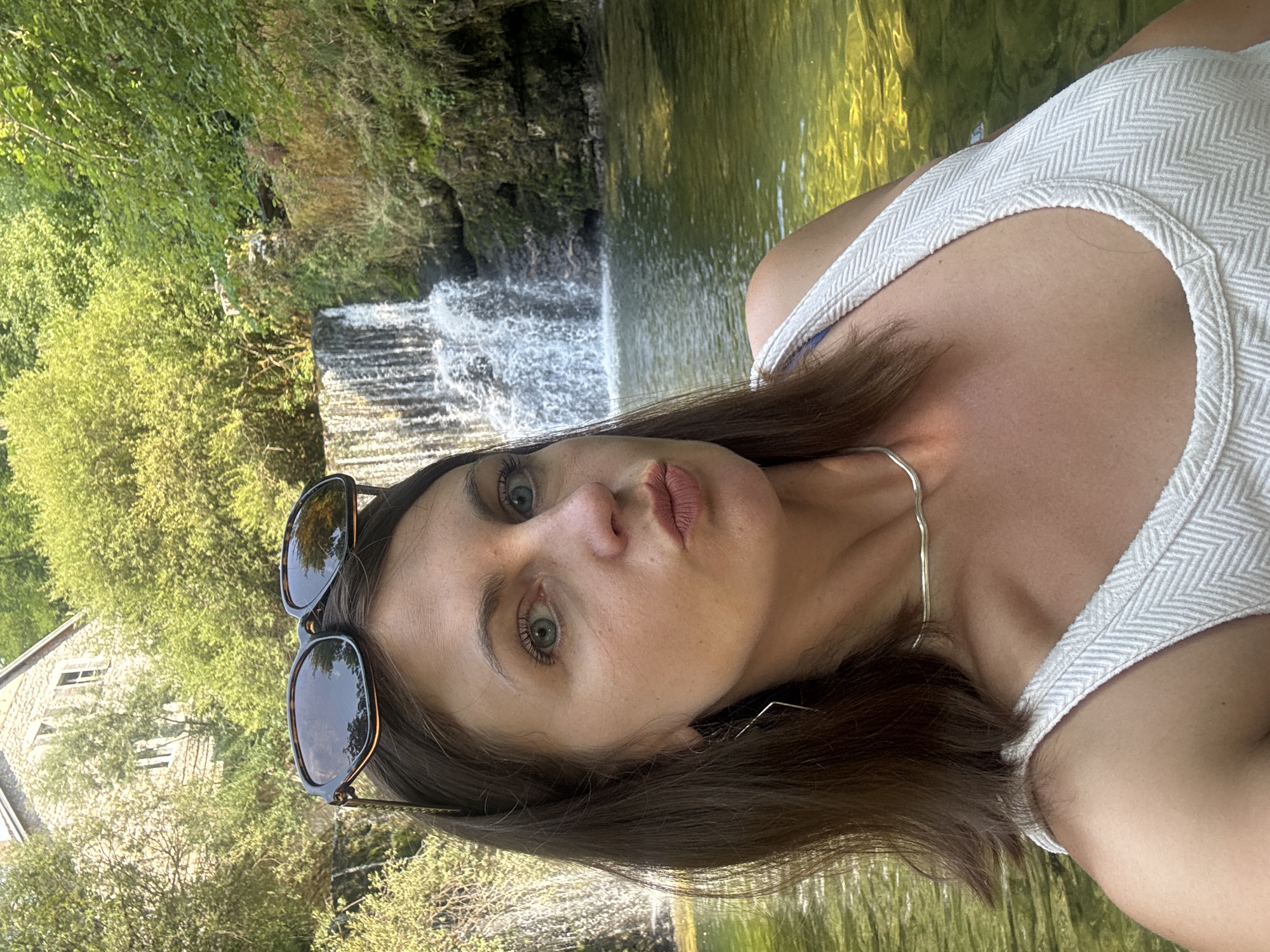 The image is a vertical, close-up selfie of a woman taken outdoors. The woman has long, brown hair and is wearing sunglasses. She is looking towards the camera with a neutral expression. She is wearing a white tank top with a subtle chevron pattern and a thin silver necklace.
The background features a waterfall cascading over rocks, surrounded by lush green trees and vegetation. The water appears clear and reflects the sunlight. To the left of the waterfall, a stone building with windows is partially visible among the trees. The overall setting suggests a natural, scenic location, possibly a park or a rural area.
The lighting indicates it is daytime, likely late afternoon given the warm tones of the sunlight. The weather appears to be pleasant and sunny. The image is cropped to focus on the woman's face and upper body, with the natural scenery serving as a backdrop. There is no visible text in the image. The location context provided is Saint-Claude, France.