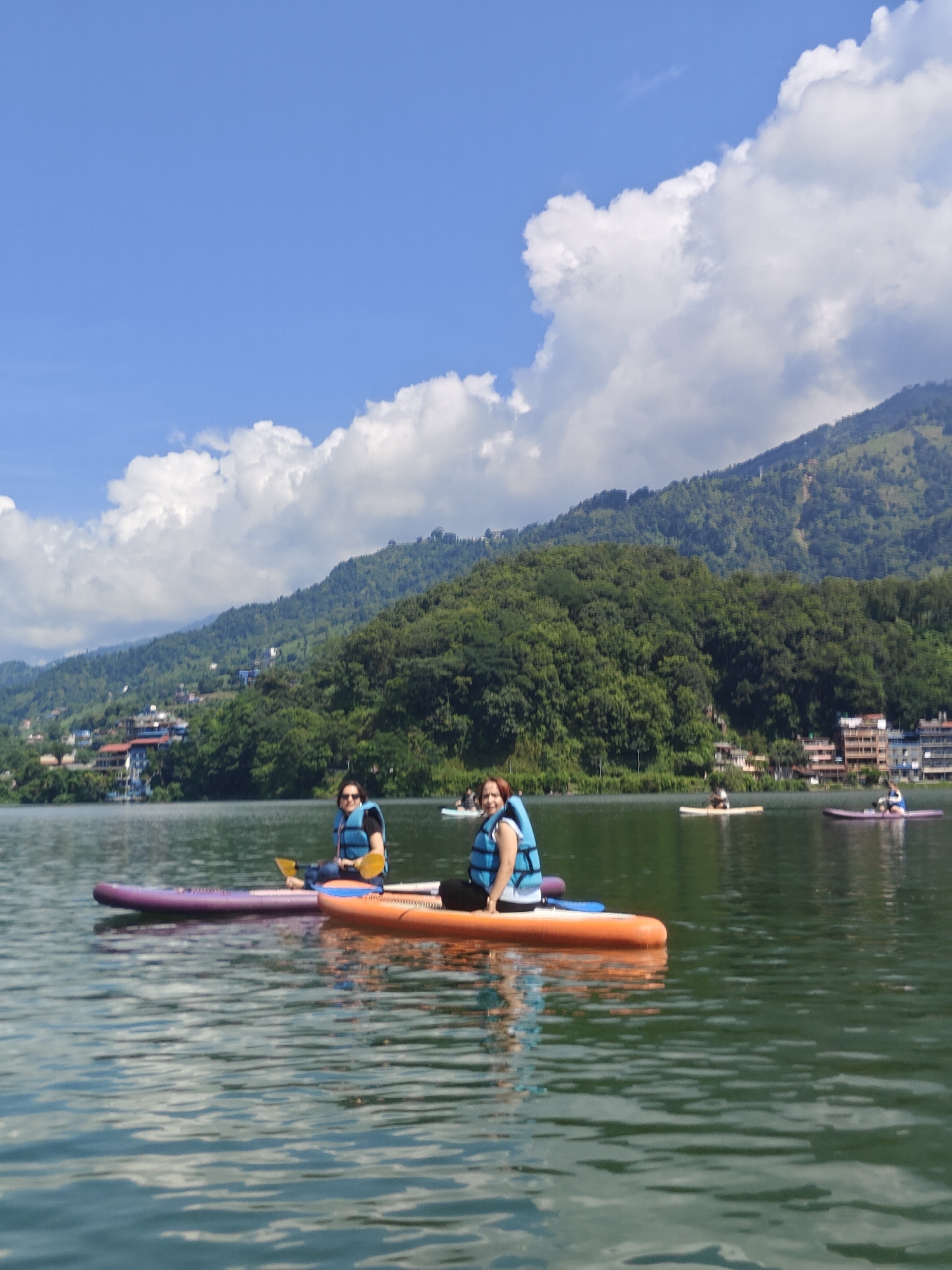 This is a ground-level, eye-level, medium shot photograph taken on a sunny day on the Phewa Lake in Pokhara, Nepal. Two women are visible, paddleboarding. The woman on the left is on a purple paddleboard, wearing a blue life vest and jeans. The woman on the right is on an orange paddleboard, also wearing a blue life vest and jeans. She is holding a yellow paddle. Both women are facing towards the camera. In the background, there are trees and buildings on a hillside, and a mountain peak is visible in the distance. The sky is blue with white clouds. Several other people on paddleboards are visible further out on the lake, as well as buildings on the shore. The water is green and reflects the sky and surrounding landscape.