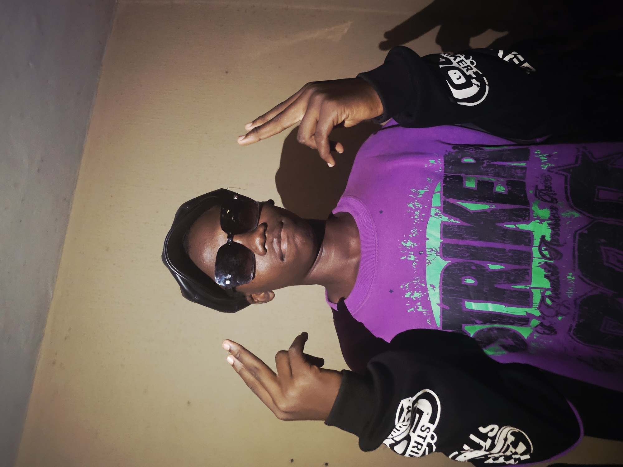 A young Black man wearing sunglasses and a black beret is pictured indoors against a beige wall. He is wearing a purple t-shirt with black and green graphics, and a black long-sleeved shirt with white graphic designs on the sleeves. He is making a hand gesture with both hands. The location is presumed to be in Nkwelle, Nigeria.
