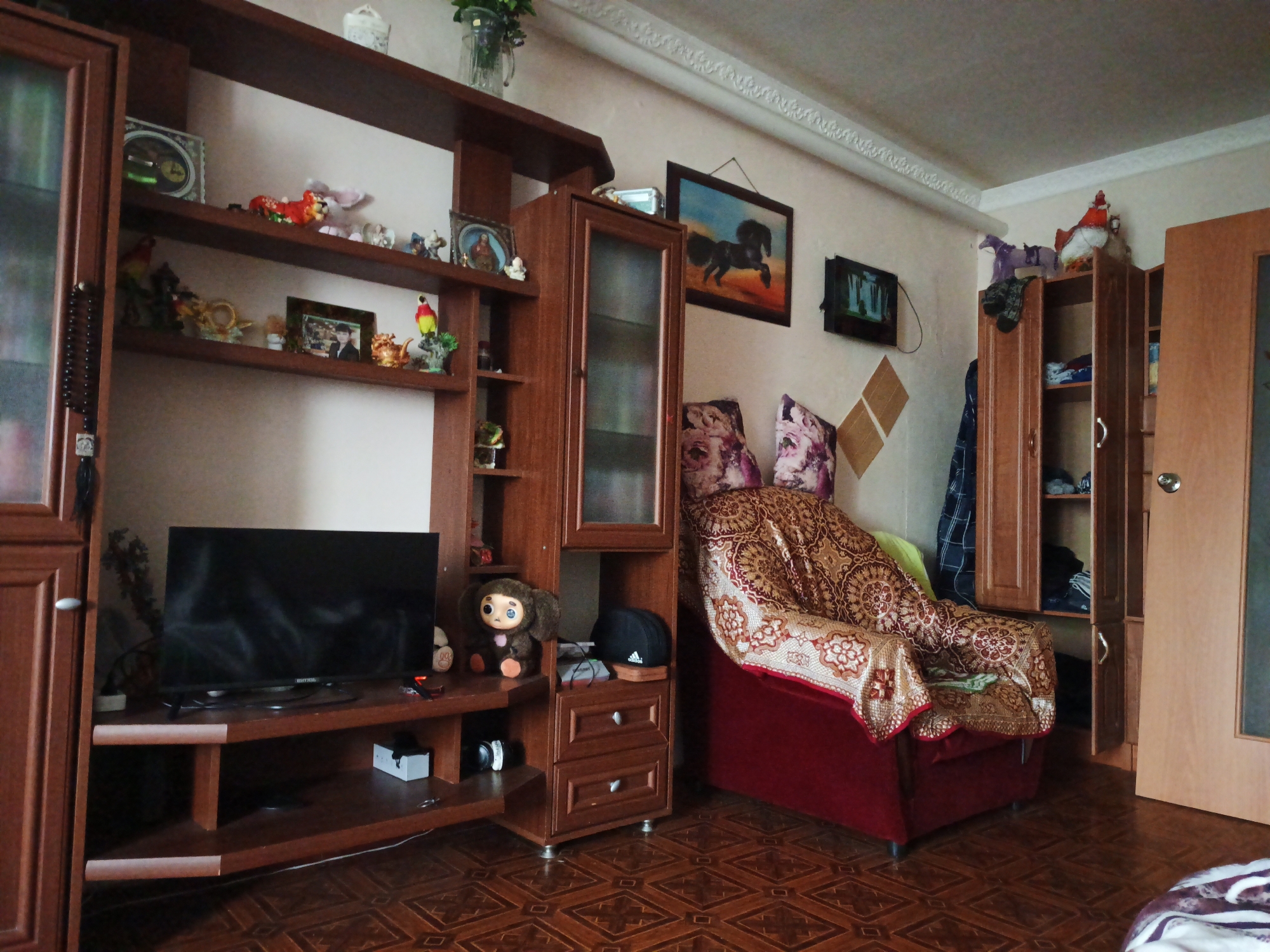 The image depicts a living room with wooden furniture and a patterned floor. A large, dark wood shelving unit with glass doors and open shelves occupies the left side of the frame. It holds various decorative items, including figurines, framed pictures, and a plush toy resembling Cheburashka. A flat-screen television is positioned on a lower shelf of this unit. To the right of the shelving unit is a red armchair draped with a patterned throw. Behind the armchair, a tall wooden cabinet with glass doors stands open, revealing shelves with stored items. A wooden door is visible to the far right of the image. The walls are a light color, and a decorative ceiling molding is present. The floor is covered with a brown, patterned linoleum. The scene appears to be a domestic interior, possibly located in Beryozovo, Russia, given the context. There are no people visible in the image.