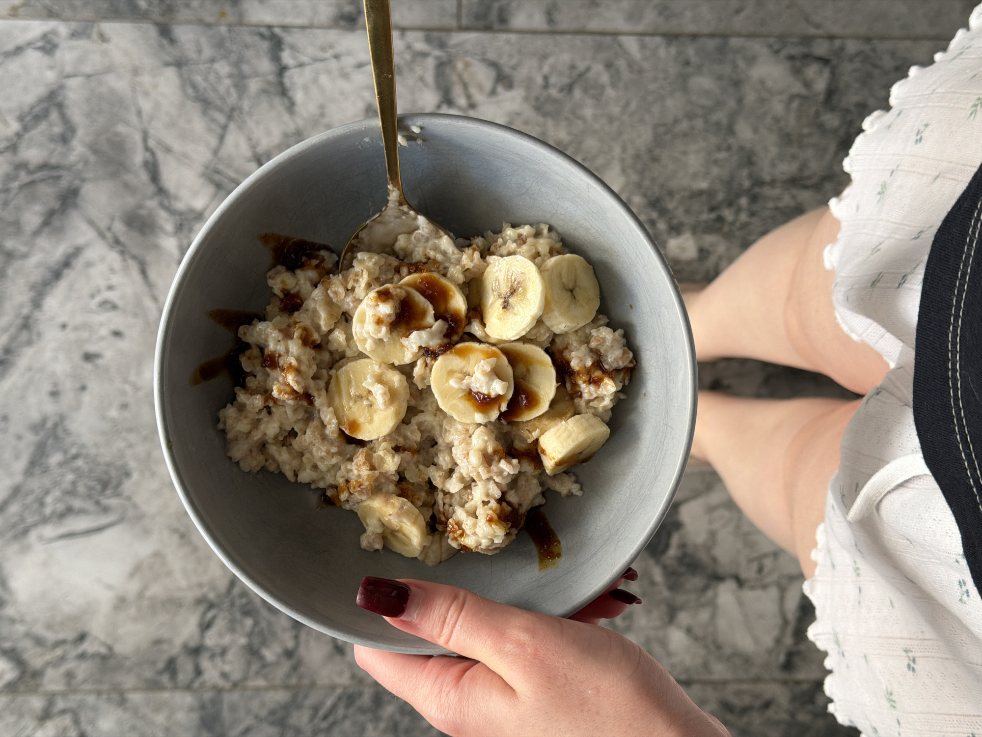 A top-down, eye-level close-up shot shows a person holding a bowl of oatmeal topped with banana slices and a dark sauce. The bowl is grey and contains a golden spoon. The person's left hand, with dark red fingernails, supports the bowl. The person is wearing a white dress with blue floral patterns and a black trim. The background is a grey, marbled surface. No information suggests that the image was taken in Rossiyskiy, Russia.