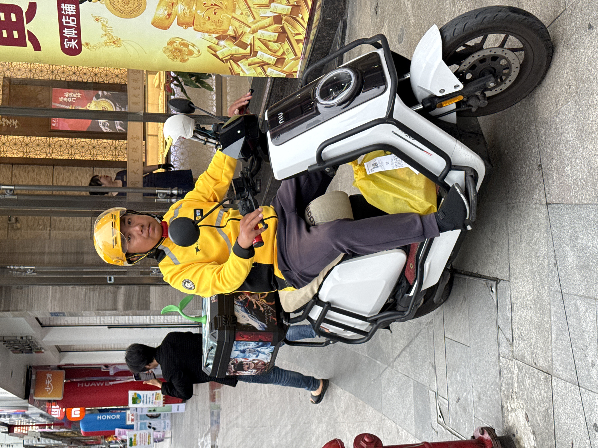 The image captures a delivery driver on a white "Niu" electric scooter in Jiangjin, China. The driver is wearing a yellow jacket and helmet and holding an item in his left hand. He is positioned on the scooter in front of a storefront with gold-themed advertisements. Behind the scooter, a person in a black shirt and jeans is looking at their phone. The scooter has a delivery container attached to the back, featuring artwork on its sides. Shop signs are visible in the background including Huawei, and Honor. The scene is set on a sidewalk paved with grey tiles.