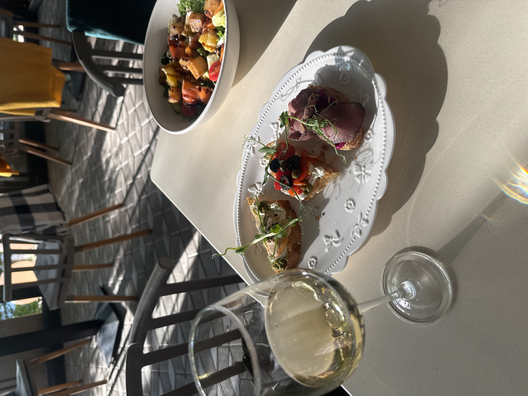 An outdoor dining area in Khabarovsk, Russia, is depicted in this image. A table is set with a glass of white wine and a white plate with three crostini. To the right of the plate, a white bowl holds a salad. In the background, chairs with cushions are visible, and a floor tiled in a geometric pattern is illuminated by sunlight casting shadows. The scene appears to be in a restaurant or cafe.