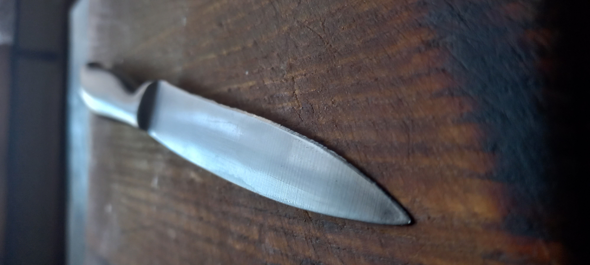 The image shows a close-up, overhead shot of a stainless steel knife lying diagonally on a rustic, textured wooden surface. The knife's blade is the focal point, reflecting light and showing subtle striations from its sharpening. The handle is partially visible on the left, appearing to be made of a smooth, lighter material, possibly plastic or metal. The wooden surface has a warm, brown tone with visible grain patterns and slight imperfections, suggesting it might be a cutting board or a well-worn table. The lighting creates a soft shadow and highlights the reflective surface of the knife, giving it a polished appearance. The background on the far left is dark and out of focus, with hints of a vertical edge. The right side of the image fades into darkness, with the wooden texture becoming less distinct. There is no visible text in the image. The scene suggests a still life composition, possibly in a kitchen or workshop setting, with no human presence. The overall mood is one of simple utility and craftsmanship.