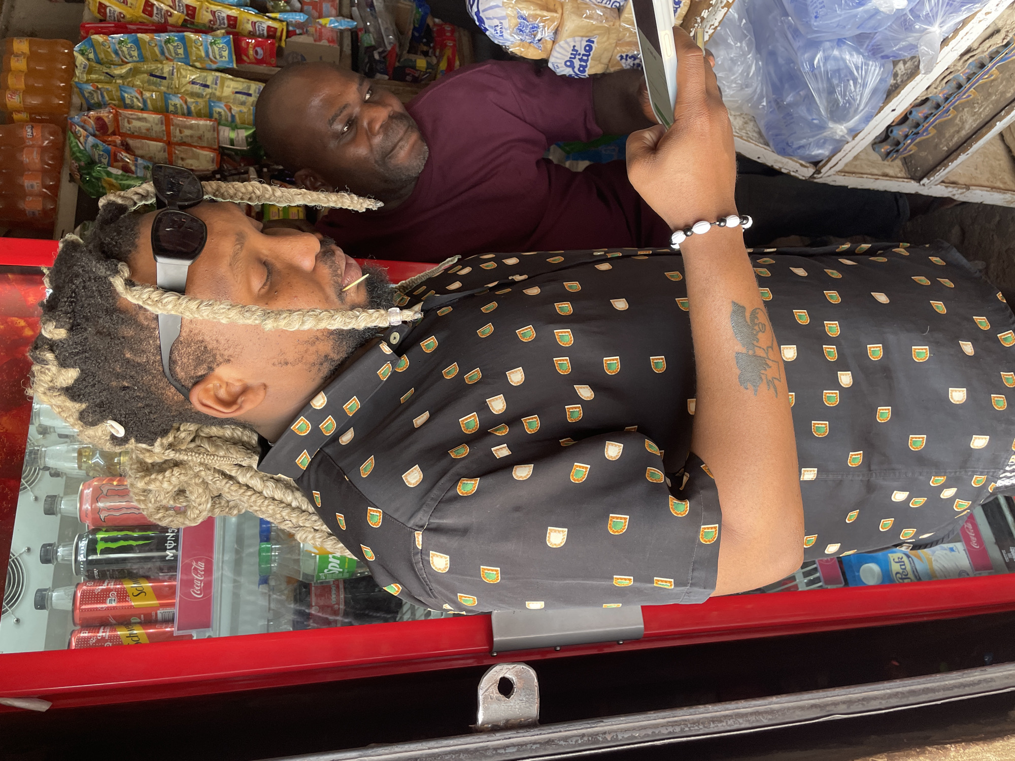 The image shows two men in what appears to be a small convenience store in Jos, Nigeria. The man in the foreground, wearing sunglasses and a patterned black shirt, is looking at a phone held in his right hand. He has blond braided hair. His left arm has a tattoo, and he wears a beaded bracelet. A second man, in a burgundy shirt, is visible in the background. The store is well-stocked with items such as bottled drinks, snacks, and bread. The setting suggests a typical urban scene in Jos, Nigeria. The image is oriented sideways, requiring rotation to view correctly.