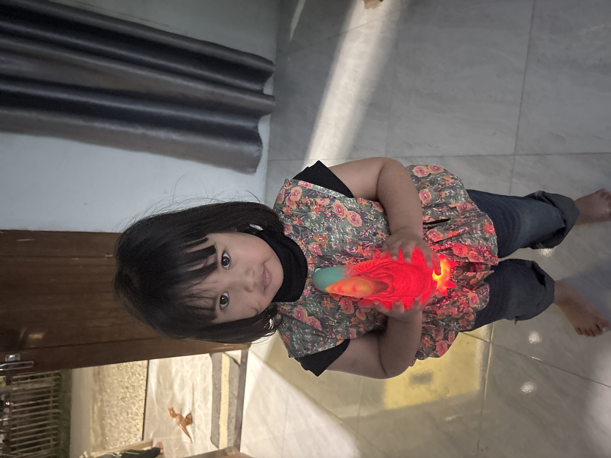 A young Indonesian girl stands in what appears to be the entranceway of a home in Lhokseumawe, Indonesia, holding a brightly lit red toy. She is wearing a black turtleneck under a floral-patterned vest and blue jeans. Her dark hair is cut short. The floor is tiled, and a doorway with a vertical wooden panel is visible to her left, leading to an outdoor area with a gravel path and a fence. To her right, a dark curtain hangs on the wall. The lighting suggests it is evening or indoors with artificial light.