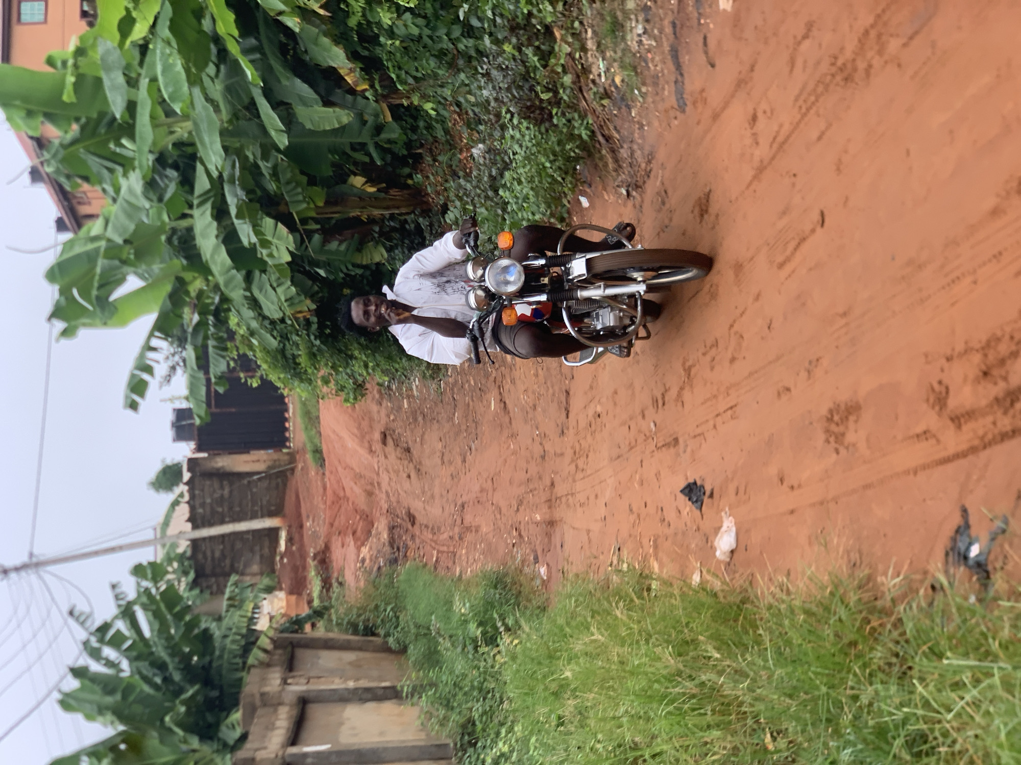 A man is seated on a motorcycle on a dirt road. He is wearing a white long-sleeved shirt and dark shorts. He is looking towards the camera and appears to be holding something up to his mouth. The motorcycle is a vintage model with chrome accents and orange turn signals. The road is made of reddish-brown dirt and has tire tracks. To the left of the road are lush green banana trees and other vegetation. In the background, there are some buildings and fences. The sky is overcast, suggesting it might be a cloudy day or early morning/late evening. The overall scene appears to be in a rural or semi-rural setting in Ukwu-Abwa, Nigeria. The man's expression is relaxed, and he seems to be enjoying himself. There is no visible text in the image.