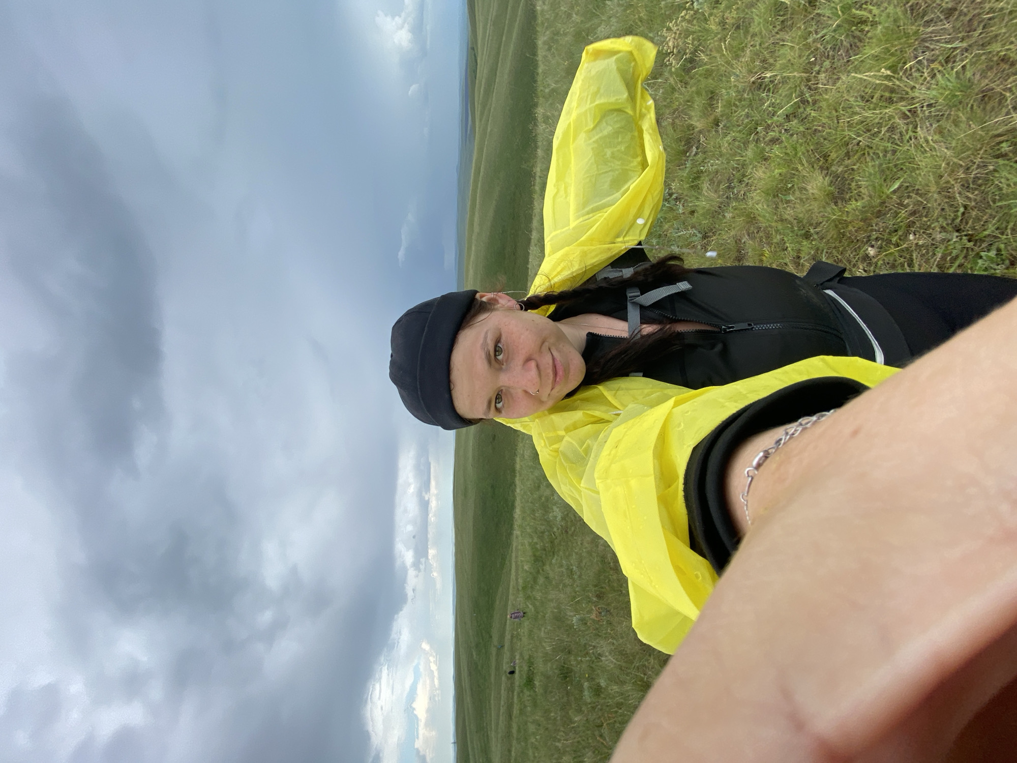 The image shows a woman in a yellow rain jacket taking a selfie in a grassy, hilly landscape under a cloudy sky. She is wearing a black beanie and has her hair in braids. The background shows rolling green hills that recede into the distance. Several other people are visible as small figures in the middle ground of the image, also in a vast, open grassy area. The location is identified as Verkhneozernoe, Russia. The overall scene suggests an outdoor activity, possibly hiking or traveling, during overcast weather.