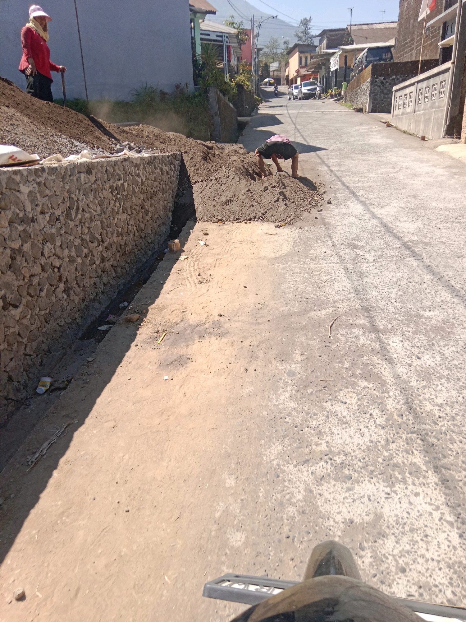 In Jurangjeru, Indonesia, a construction project is underway on a street. A person wearing a red long-sleeved shirt, black pants, and a pink hat is standing on a pile of dirt, holding a shovel. Dust is being kicked up around them. Another person, wearing dark shorts and a striped shirt, is crouched down next to a pile of dirt on the street, appearing to be working. The street is paved and slopes uphill, with several buildings visible on either side. A stone wall has been constructed alongside the road, with a narrow drainage ditch running next to it. The scene suggests ongoing manual labor and infrastructure work in a residential or semi-rural area.