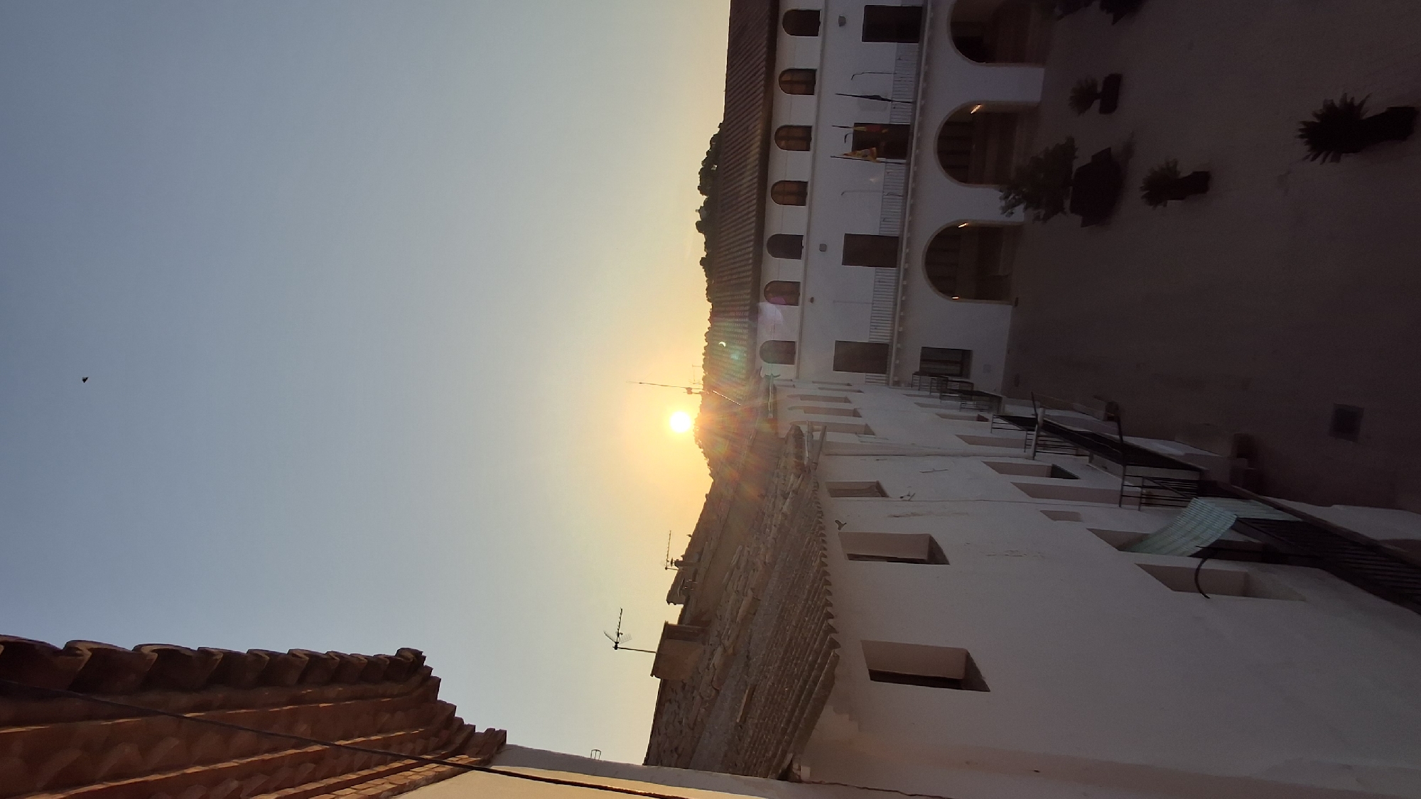 The image captures a courtyard view of buildings in Híjar, Spain, during sunset. A clear, pale blue sky dominates the upper portion of the frame, with the sun appearing as a bright, orange orb setting behind a distant, tree-covered hill. Sunbeams emanate from the sun, creating a lens flare effect.
The foreground features the tiled roof of a building on the left, with its white exterior wall and rectangular windows visible. The wall appears to be aged, with some texture. Extending from the building is a balcony with black wrought-iron railings.
To the right, a larger building forms the side of the courtyard. This building has a white facade with several arched openings on the ground floor and rows of arched windows on the upper level. Flags hang from poles on the upper floor of this building, displaying red and yellow colors. Balconies with similar black railings are present on this structure as well.
The courtyard floor is paved with a light-colored material, and several potted plants are scattered throughout, adding greenery to the scene. The lighting suggests it is late afternoon or early evening, with shadows beginning to lengthen across the courtyard. A small, dark object, possibly a bird or an insect, is visible in the sky near the top center of the image.