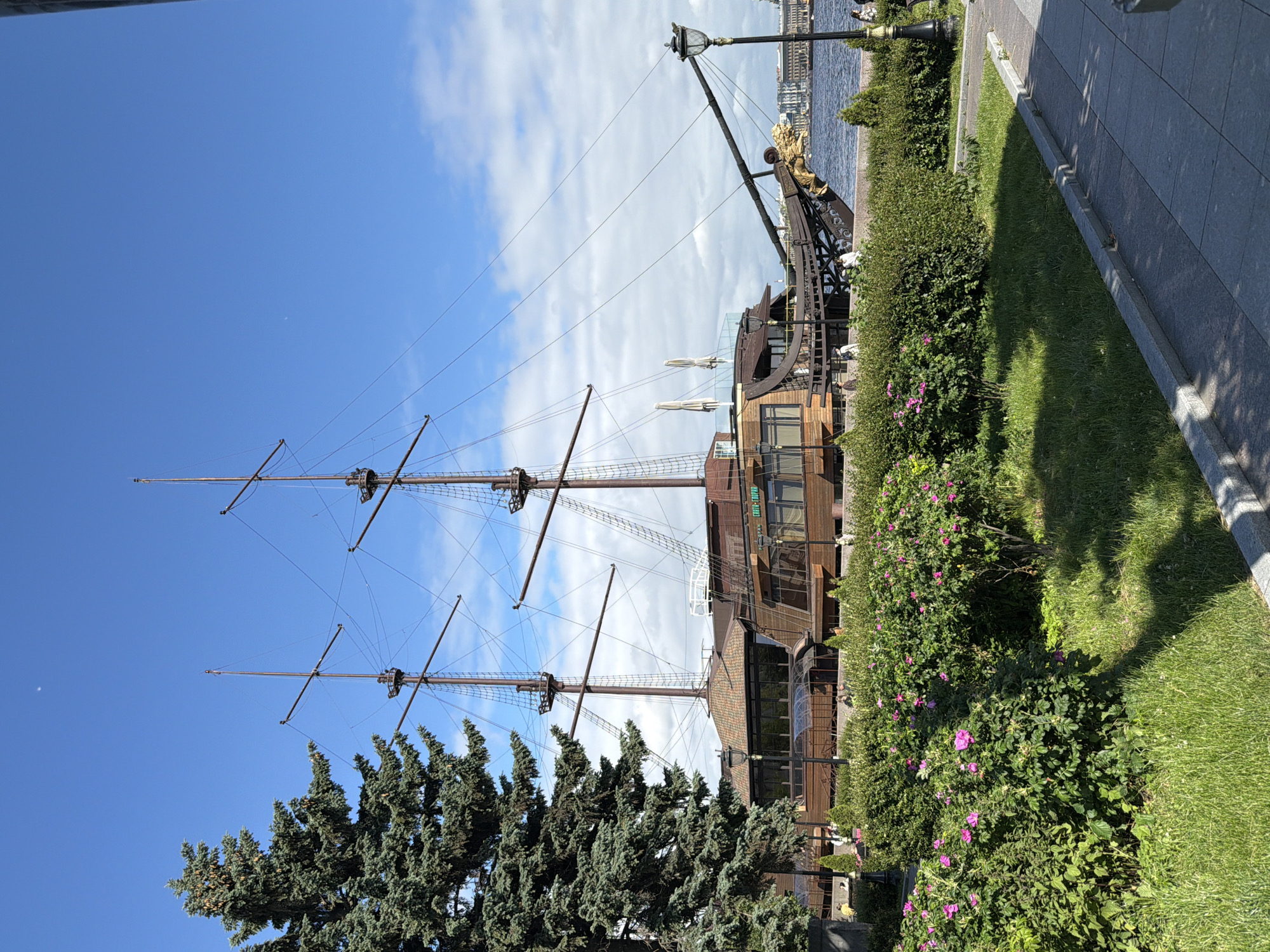 This is a vertical, eye-level, full-shot photograph of a docked, wooden, tall ship restaurant with a golden dragon figurehead. The ship has two masts and rigging. The ship is brown and has many windows. In front of the ship is a green lawn with rose bushes in bloom. Behind the ship is a river and a city skyline with several buildings. The sky is blue with some white clouds. The photograph was taken on a sunny day. The ship is located in Saint Petersburg, Russia.