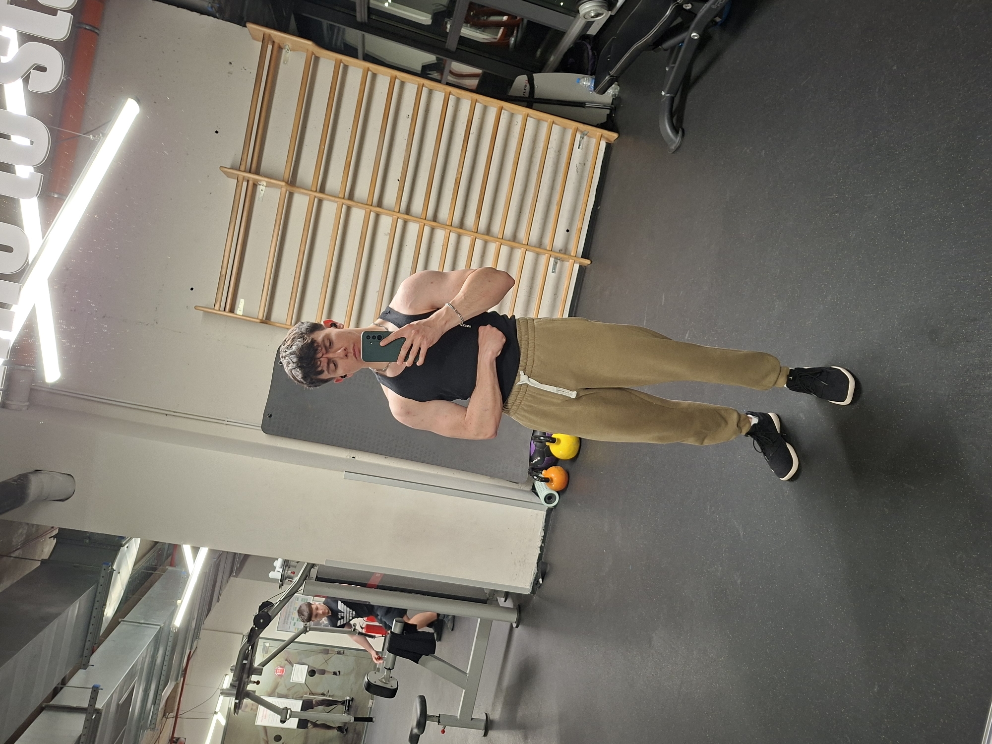 A selfie photograph depicts a young man in a gym setting. The man is wearing a black tank top and tan sweatpants with black sneakers. He is holding a cell phone in his left hand, taking a mirror selfie. In the background, there is gym equipment including wooden wall bars, weights, and weightlifting machines. Another individual is visible using the gym equipment in the background. The location appears to be a gym, but it is impossible to confirm that the location is in the city of Plovdiv, Bulgaria based on the image alone.