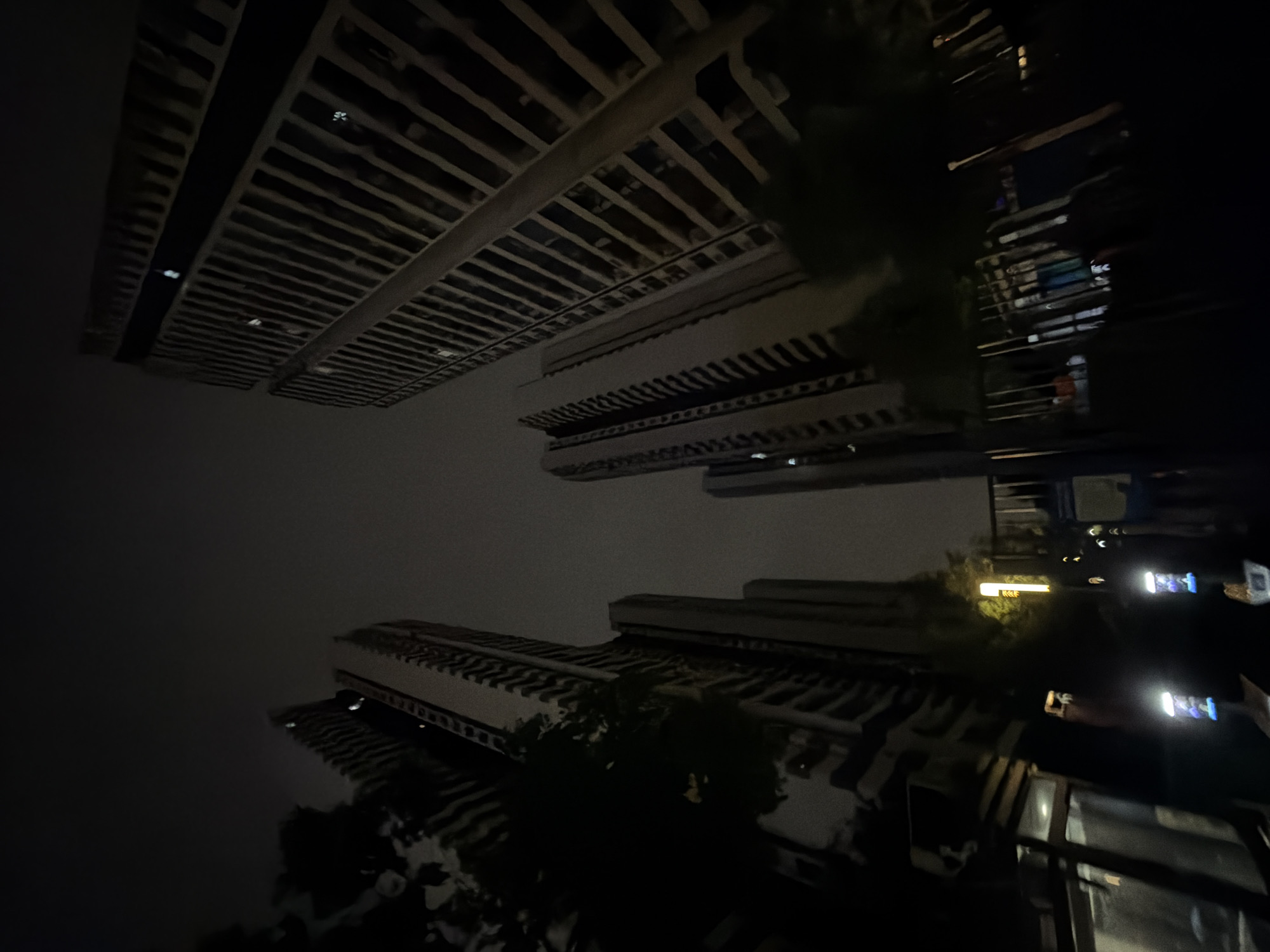 The image depicts a nighttime view of several high-rise buildings in the Chongqing High-tech Zone, China. The perspective is looking upwards from ground level, emphasizing the verticality of the structures. The buildings are tall and appear to be apartment complexes or office buildings, with many windows visible. Some windows are illuminated, suggesting occupancy. Trees are present in the foreground, casting shadows and obscuring parts of the buildings. In the lower portion of the image, there are glimpses of what appears to be a public space or park area, with railings, some indistinct objects, and several bright lights that could be streetlights or decorative lighting. The sky is dark and overcast, contributing to the nighttime ambiance. The overall scene suggests an urban environment at night.
