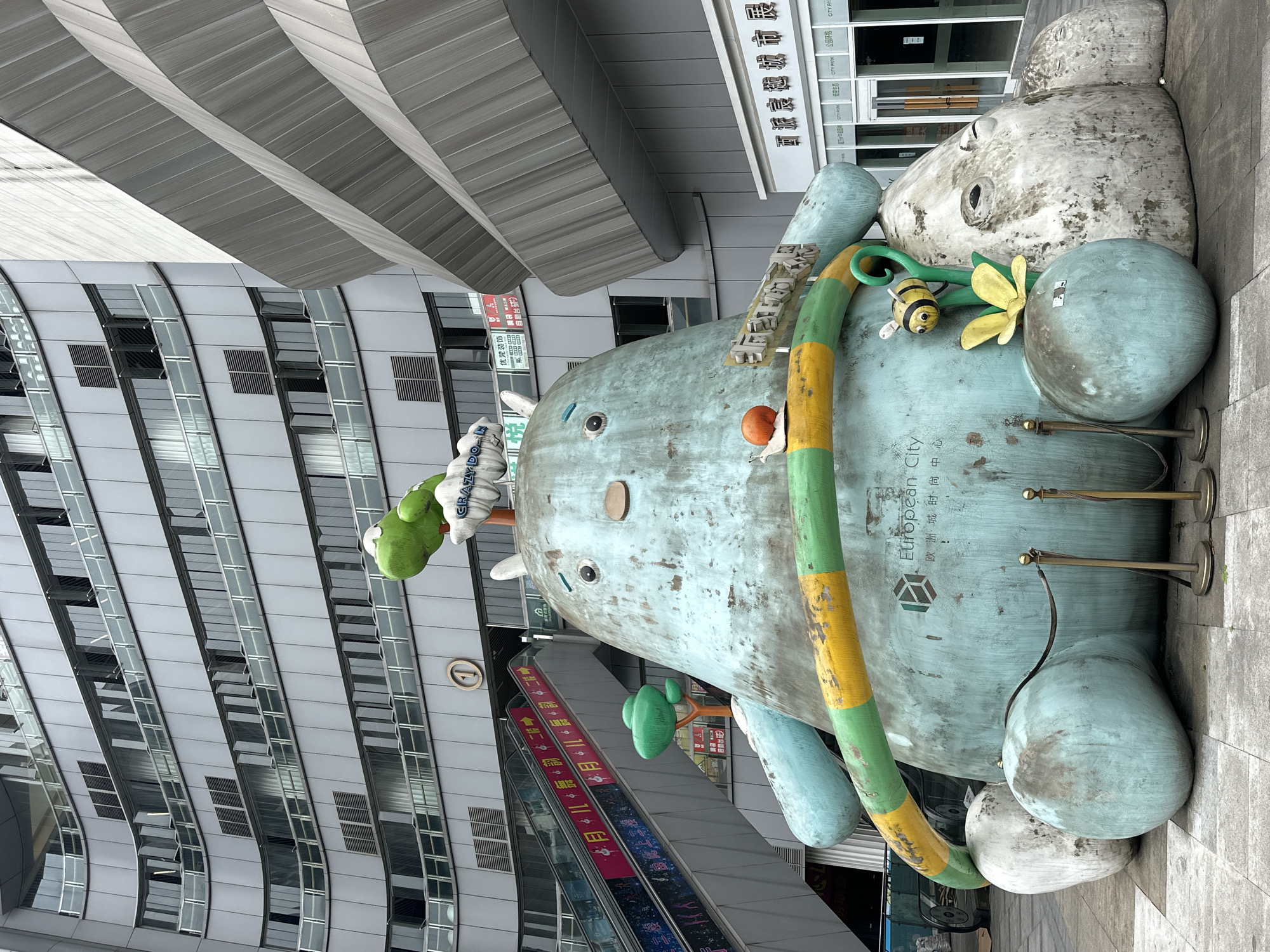 A large, blue-green, somewhat weathered sculpture of an animal, possibly a hippo or elephant, is the focal point of the image. The sculpture has cartoonish features, including a large nose, eyes, and other embellishments. It is adorned with a green and yellow striped hoop and a bee attached to a yellow flower.
The sculpture is situated outdoors, likely in a public space or plaza in Jiulongpo District, Chongqing, China. An escalator is visible to the side of the sculpture. A sign on the sculpture reads "European City" in English and Chinese characters. A modern building with a facade of glass windows rises in the background, suggesting a commercial or urban environment. The image is taken at ground level.