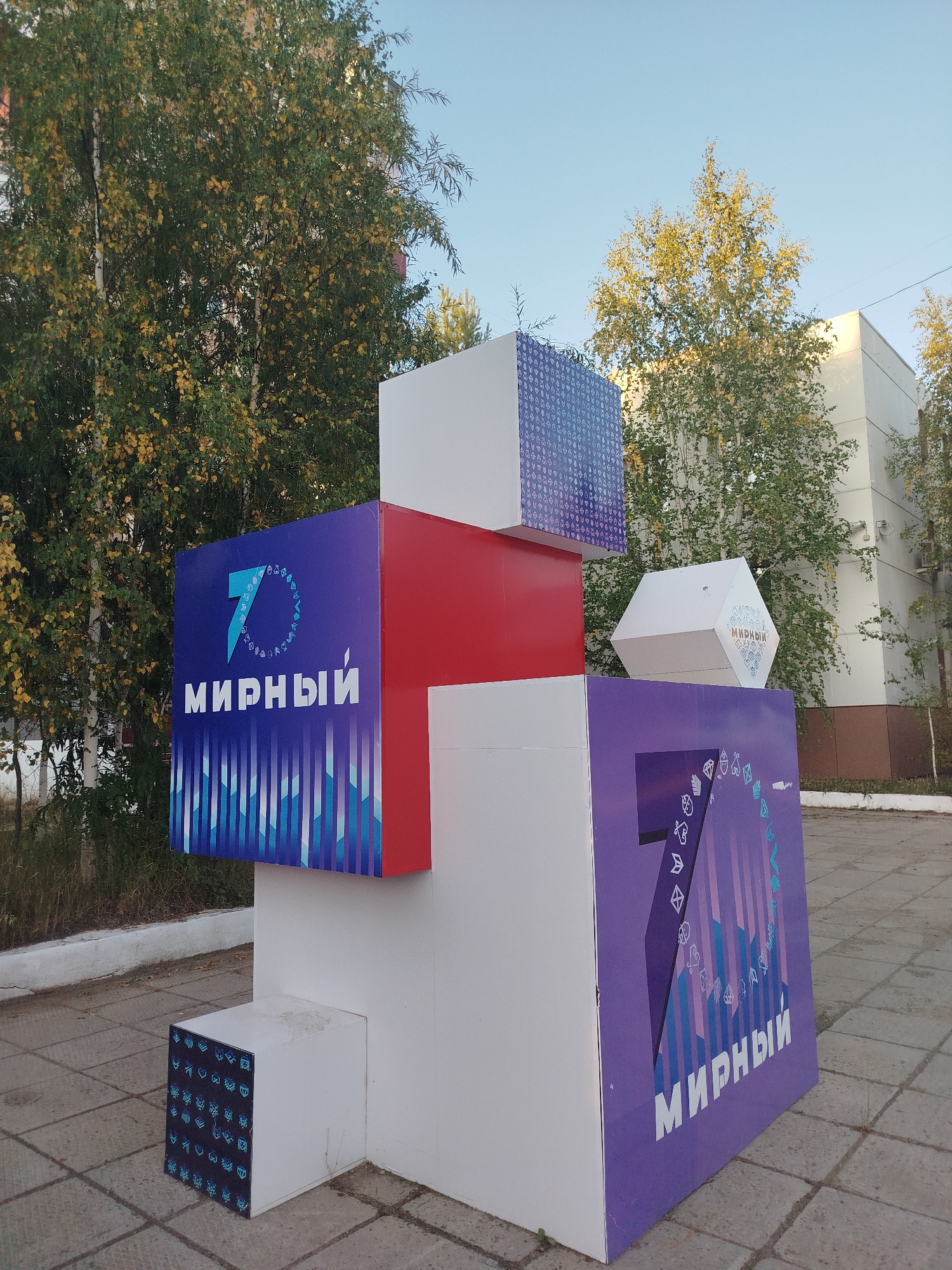 The image shows a large, multi-faceted art installation located in the city of Mirny, Russia. The installation is composed of several rectangular and hexagonal blocks, predominantly white, with sections adorned with blue, purple, and red panels featuring geometric patterns and text. The text "МИРНЫЙ" (Mirny) is visible on at least two of the panels, along with the number "7" and stylized designs. The installation is situated on a paved area, likely a public space or plaza, with trees and buildings visible in the background. The sky is clear and blue. There are no people or animals present in the image. The overall scene depicts an outdoor urban environment.
