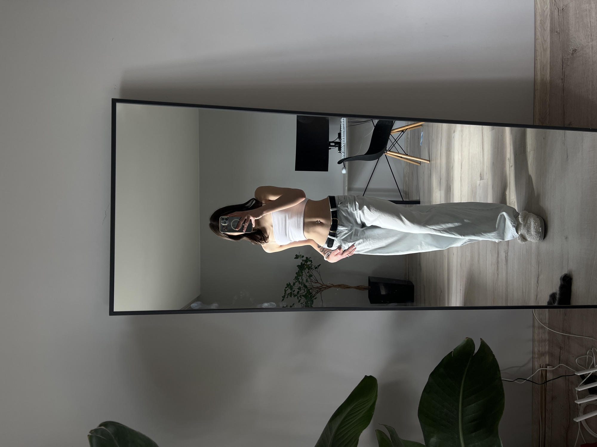 A person is standing in front of a full-length mirror in a room. The person is wearing a white strapless top, light-colored baggy pants, a black belt, and fuzzy slippers. They are holding a smartphone up to take a photo of themselves. In the reflection of the mirror, a desk with a computer monitor and a chair can be seen. To the left of the mirror, a large plant is visible. The room has light gray walls and a light wooden floor. The lighting suggests it is daytime, with sunlight casting shadows. This scene is not in Ternopil, Ukraine, as no specific location details are provided in the image.