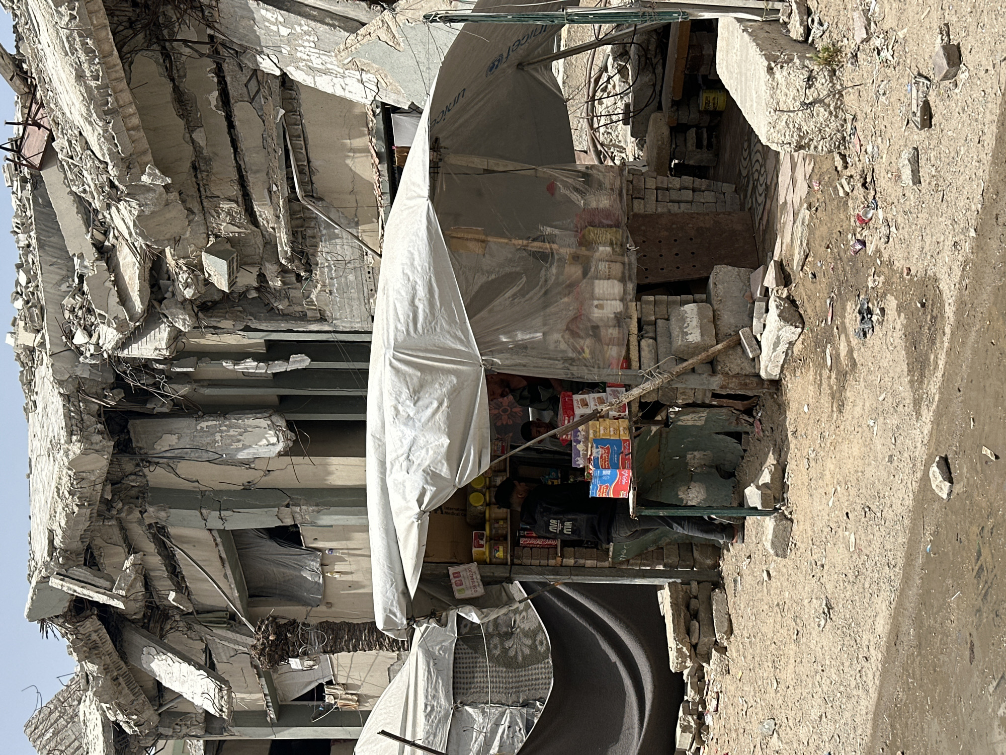 The image depicts a damaged building, likely in Gaza City, Palestinian Territories, given the presence of a UNICEF-branded shelter. The building is severely damaged, with exposed concrete and rebar. A makeshift shop has been erected in front of the ruins, selling various goods. A person is standing in the shop. The surrounding ground is littered with debris.