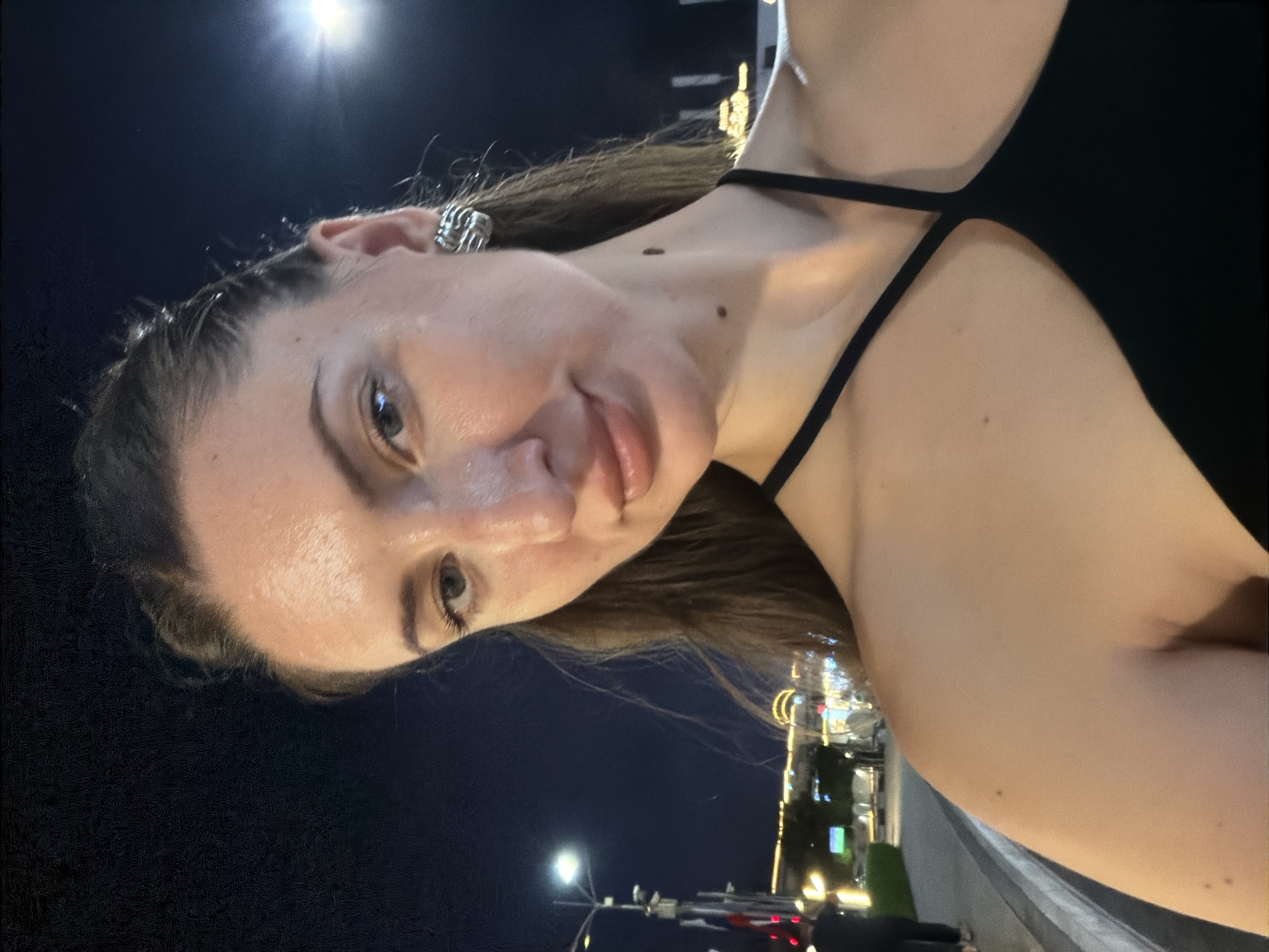 A young woman with long brown hair and fair skin is pictured at night in Sharm El Sheikh, Egypt. She is wearing a black halter top and large, silver, circular earrings. The woman is looking at the camera with a slight smile. In the background, there are streetlights and buildings adorned with festive lights. A street runs along the left side of the image, with a person walking in the distance. The sky is dark blue, indicating it is nighttime.