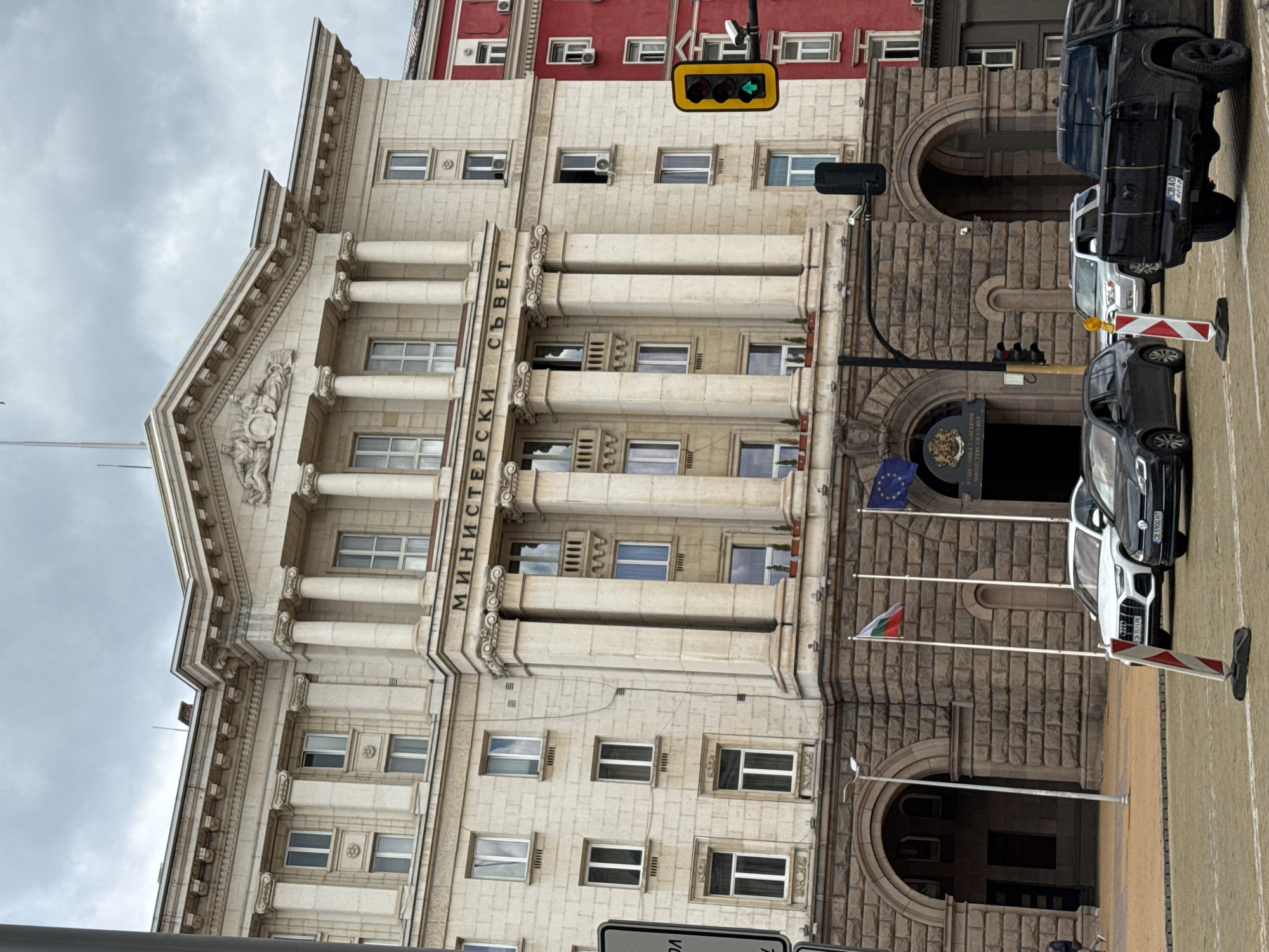 The image captures the architectural grandeur of the Council of Ministers building in Sofia, Bulgaria. The structure is an imposing edifice with a classical design featuring columns, intricate moldings, and the inscribed Cyrillic text "МИНИСТЕРСКИ СЪВЕТ" (Council of Ministers). The facade combines both light-colored stone or concrete with darker, roughly textured masonry at the lower levels.
To the right of the building, vehicles are parked or moving, including a black SUV with license plate CBAO 4050. A traffic light is visible, showing a green arrow, and there's also a road marker in the foreground. The flags of Bulgaria and the European Union fly near the entrance. The sky is overcast, suggesting a cloudy day in Sofia. The presence of modern elements such as air conditioning units on the adjacent red building juxtaposes the classic architecture. The image also captures a pedestrian walkway or sidewalk in the foreground.