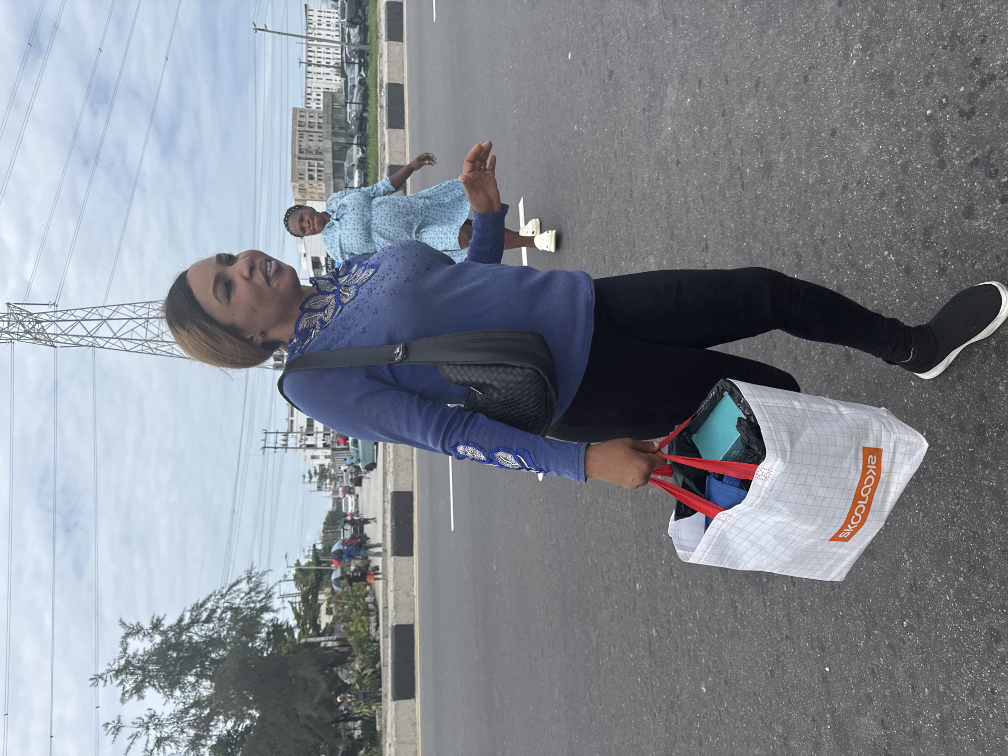 This image, captured in Igboefon, Nigeria, features two women on a paved road under a bright, partly cloudy sky. The primary subject, positioned in the foreground and oriented to the right, is a woman with light brown hair. She is wearing a long-sleeved blue top adorned with white embroidered patterns on the chest and sleeves, black leggings, and black and white sneakers. A black shoulder bag is slung across her body, and she holds a white shopping bag with red handles in her right hand. The shopping bag contains a blue box and other items, and the brand "SKOOLOOKS" is visible in red text on its side. Her facial expression is pleasant, with a slight smile, as she looks upwards and to the left.
Further in the background, another woman is seen walking or standing on the road. She is dressed in a light blue or white patterned dress and white shoes. The scene is set on a grey asphalt road with black and white curbs, suggesting an urban or semi-urban environment. To the left, a large metal power pylon with numerous power lines extends skyward. In the distance, along both sides of the road, various buildings, some appearing multi-storied, and parked vehicles are visible, along with scattered trees and greenery. The overall impression is a bright daytime scene on a street.