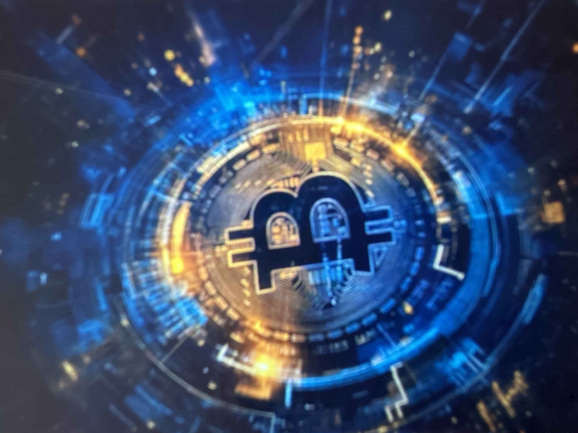The image shows a close-up, blurred view of a digital representation of Bitcoin. The Bitcoin symbol, a stylized 'B', is centrally located, surrounded by concentric circles of blue and orange light. The background consists of a dark, abstract digital landscape with glowing points of light. There is no indication of any location in the city of Gelendzhik, Russia, or any people, actions, or interactions.