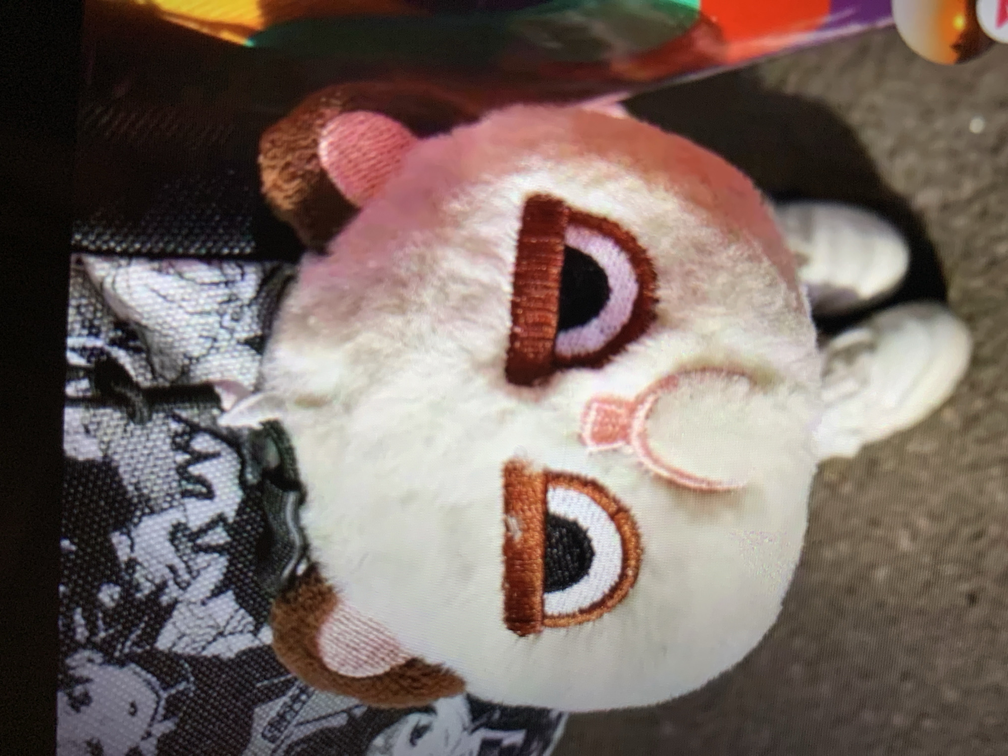 A plush toy, resembling a grumpy bear with white and brown fur, is the central subject of this image. It has large, downturned embroidered eyes with orange rims and black pupils, and a small pink embroidered nose and mouth. The toy is adorned with two small brown fuzzy ears and pink inner ear details. A black ribbon is tied around its head. The plush toy is positioned in front of a backpack or bag with a black and white anime-style print. The toy appears to be standing on a paved surface, with glimpses of white sneakers peeking out from beneath it. In the background, to the right, a colorful, possibly cylindrical object with segments of red, purple, and green is visible.
The image location is stated to be the city of Lymanka, Ukraine. However, there is no visual information within the image itself that would allow for confirmation of this specific location. The scene depicted is a close-up of the plush toy and its immediate surroundings.