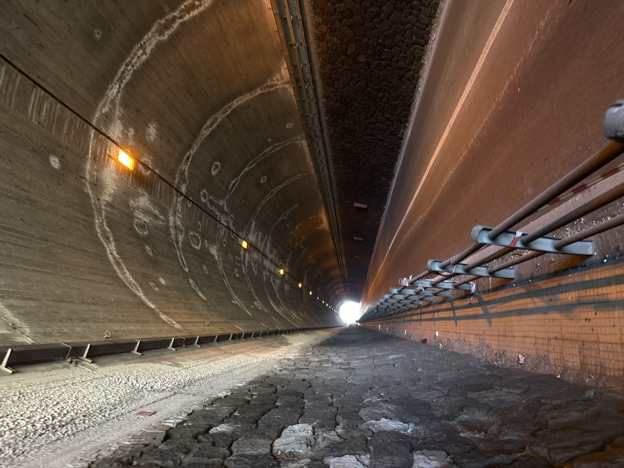 The image displays a wide-angle, low-angle perspective looking down a long, dark tunnel towards a bright light at its far end, suggesting an exit or powerful light source. The tunnel appears to be disused or unfinished, located in Lipari, Italy.
The left side of the tunnel features a tall, curved concrete wall marked by numerous large, white, circular and semicircular stains, possibly mineral deposits or water damage. Several small, rectangular, amber-orange lights are mounted on a horizontal wire running along this wall, providing sparse illumination. Below these lights, a dark horizontal beam is supported by angled white brackets, bordering a bed of light-colored gravel or small stones on the ground.
The right side of the tunnel presents a contrasting, flatter, reddish-brown wall. Higher up, a series of metal bars or pipes project horizontally from the wall, forming a ladder-like structure, some adorned with small red and white striped markings. Below this, the wall exhibits a rough, textured surface, possibly brick or stone, with dark streaks and debris near the base.
The central floor of the tunnel is uneven and dark, composed of large, irregularly shaped stones or paving blocks that resemble rough cobblestones or broken concrete. This rugged terrain extends towards the distant, bright white light. No people or visible text are present in the image. The scene conveys a sense of depth, decay, and the long journey through an enclosed space.