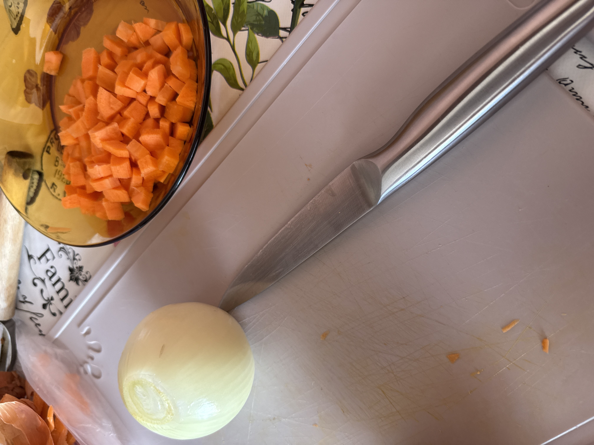 A top-down view captures food preparation in progress. A clear glass bowl contains diced carrots, positioned in the upper left corner. A peeled, whole onion lies on a white cutting board. A silver-handled knife rests diagonally across the cutting board, with its blade near the onion. Peels are also visible. The environment appears to be a kitchen or food preparation area. There is no clear indication about the city of Sofia, Bulgaria.