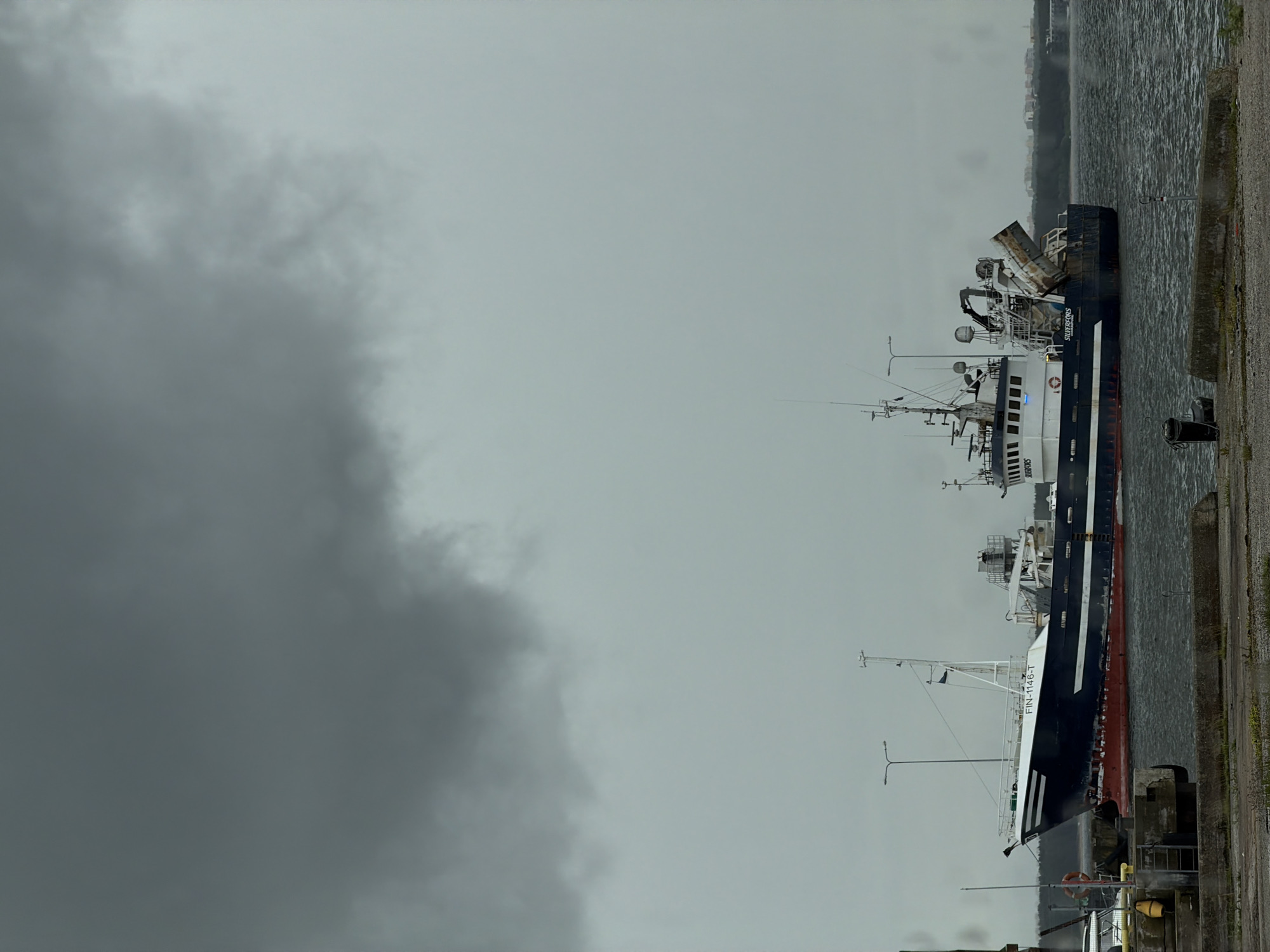 Two fishing trawlers are docked in the harbor of Viimsi, Estonia, under a cloudy, overcast sky. The larger vessel in the foreground, named "SILVERFORS" and bearing the registration "FIN-1146-T", is dark blue with a red hull and white superstructure. It is equipped with fishing gear, including a large crane and what appears to be a processing unit. The second trawler, partially visible behind the first, is also dark blue and white. In the background, a low-lying coastline with buildings is visible through the mist. The water is choppy, and the overall atmosphere is somber due to the weather. The scene appears to be captured from a concrete pier.