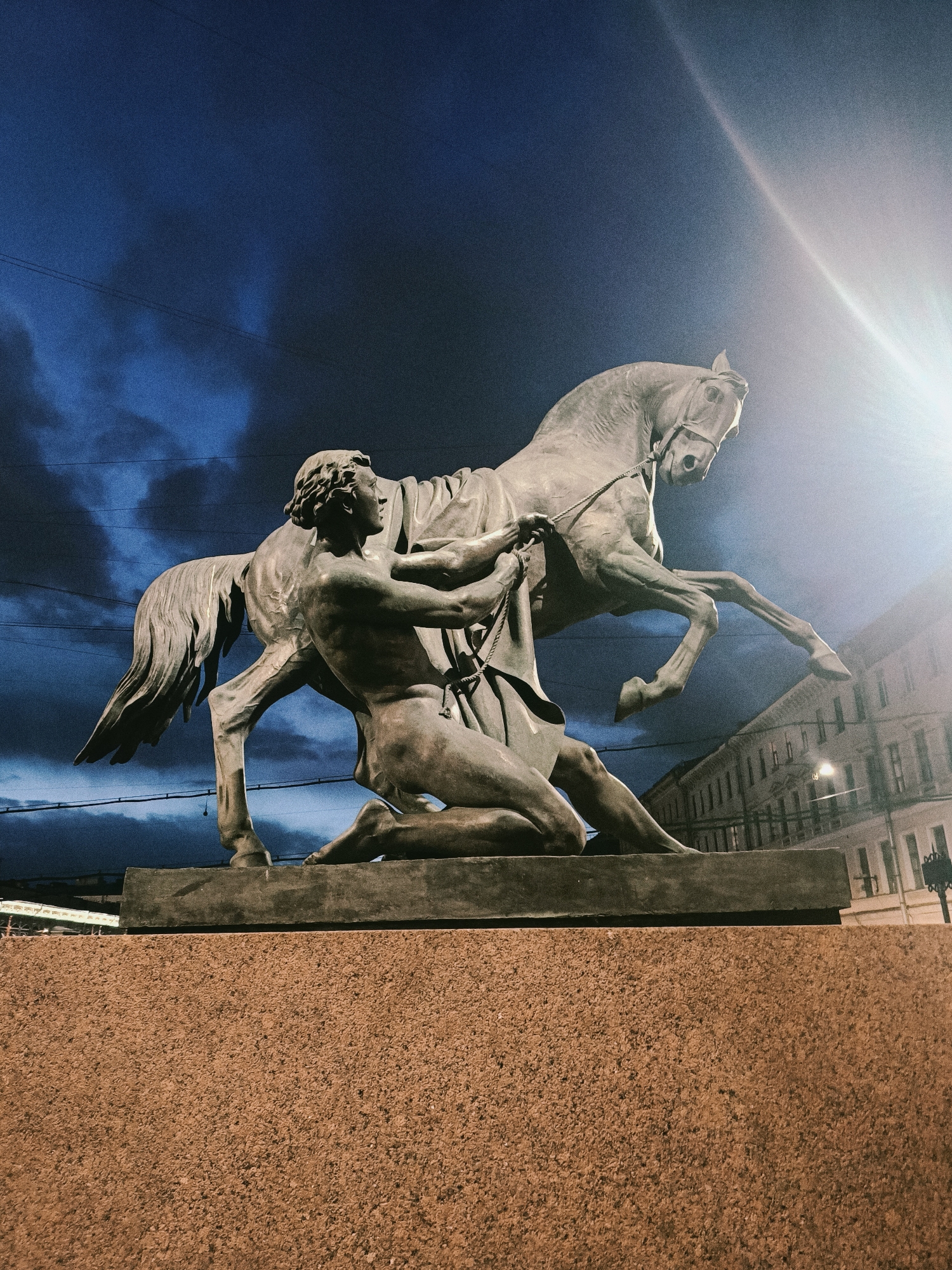 The image shows a statue depicting a nude male figure struggling to control a rearing horse. The statue is positioned on a stone pedestal. Behind the statue, the night sky is visible, with the silhouettes of buildings partially illuminated by a light source. The statue is located on the Anichkov Bridge in Saint Petersburg, Russia.
