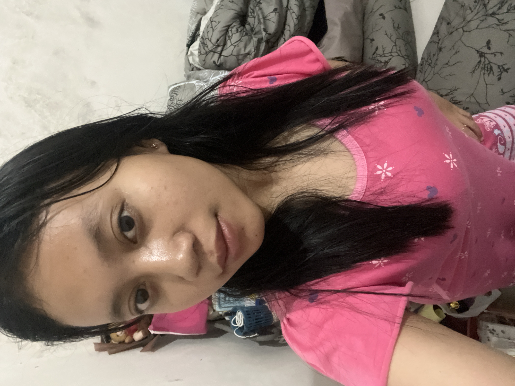 An eye-level, medium close-up selfie shows a young woman with straight, black hair framing her face and falling past her shoulders. She is wearing a bright pink t-shirt with a pattern of small white flowers and purple heart shapes. Her skin is moist, possibly from sweat or water. She has dark eyes and a neutral expression. The background is out of focus, but appears to be an indoor setting with a white wall on the left and a pile of gray and white patterned bedding on the right. A brown stuffed bear is partially visible on a shelf to the left of the woman. The scene is likely a bedroom. The provided location is Jambearum, Indonesia.