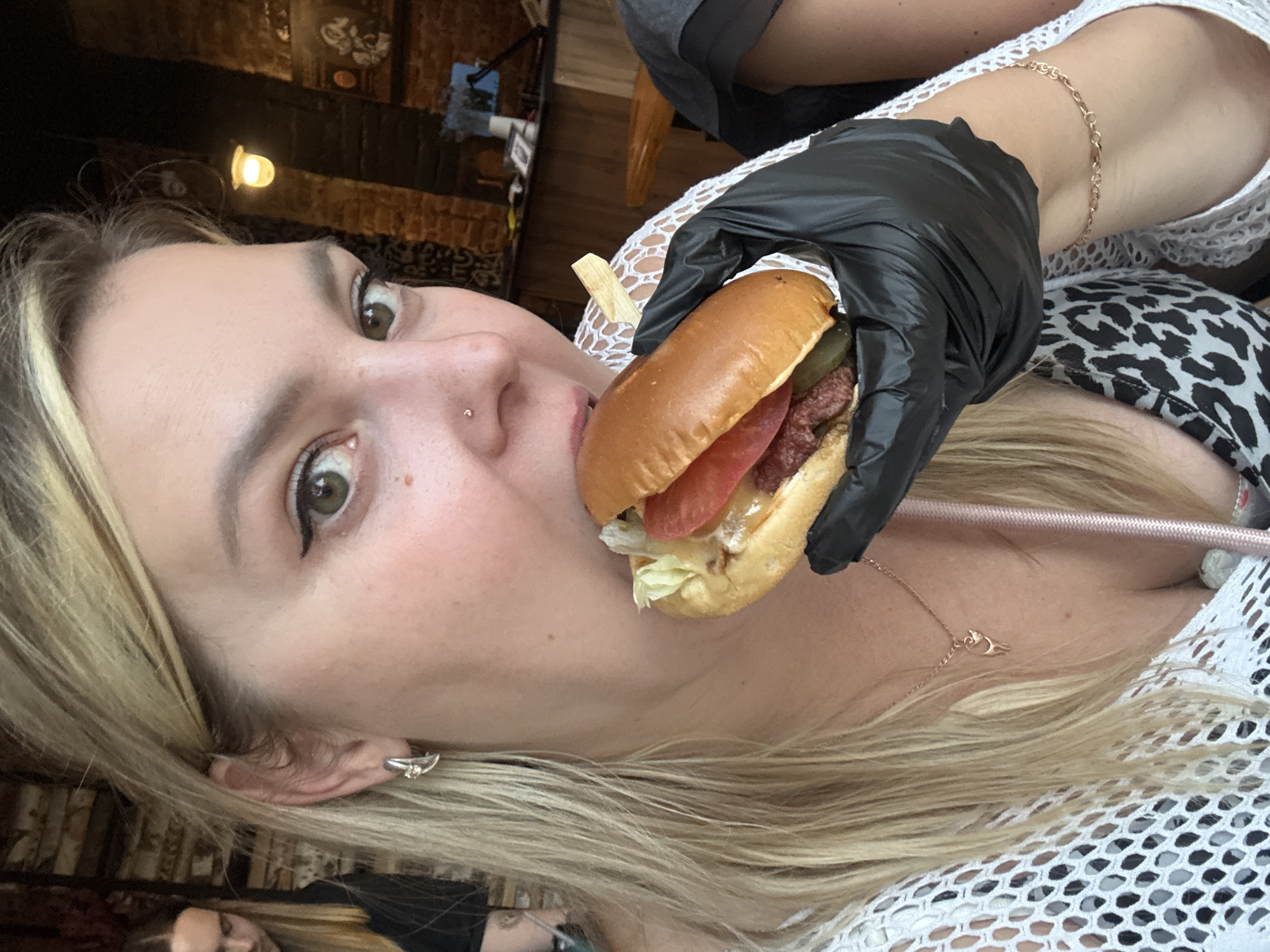 The media file shows a close-up, rotated shot of a person wearing black gloves and holding a hamburger to their mouth. The person has blonde hair, green eyes, and is wearing a white, mesh-like top. A silver earring is visible on their left ear, and a delicate gold necklace is around their neck. The background appears to be indoors, with warm lighting from overhead fixtures and exposed brick walls suggesting a restaurant or bar setting. A second person's face is visible in the lower portion of the image, reflected in a surface, and they appear to be looking down. The image is captured from a high-angle perspective, with the focus on the hamburger and the person's face. There are no visible signs of text.
